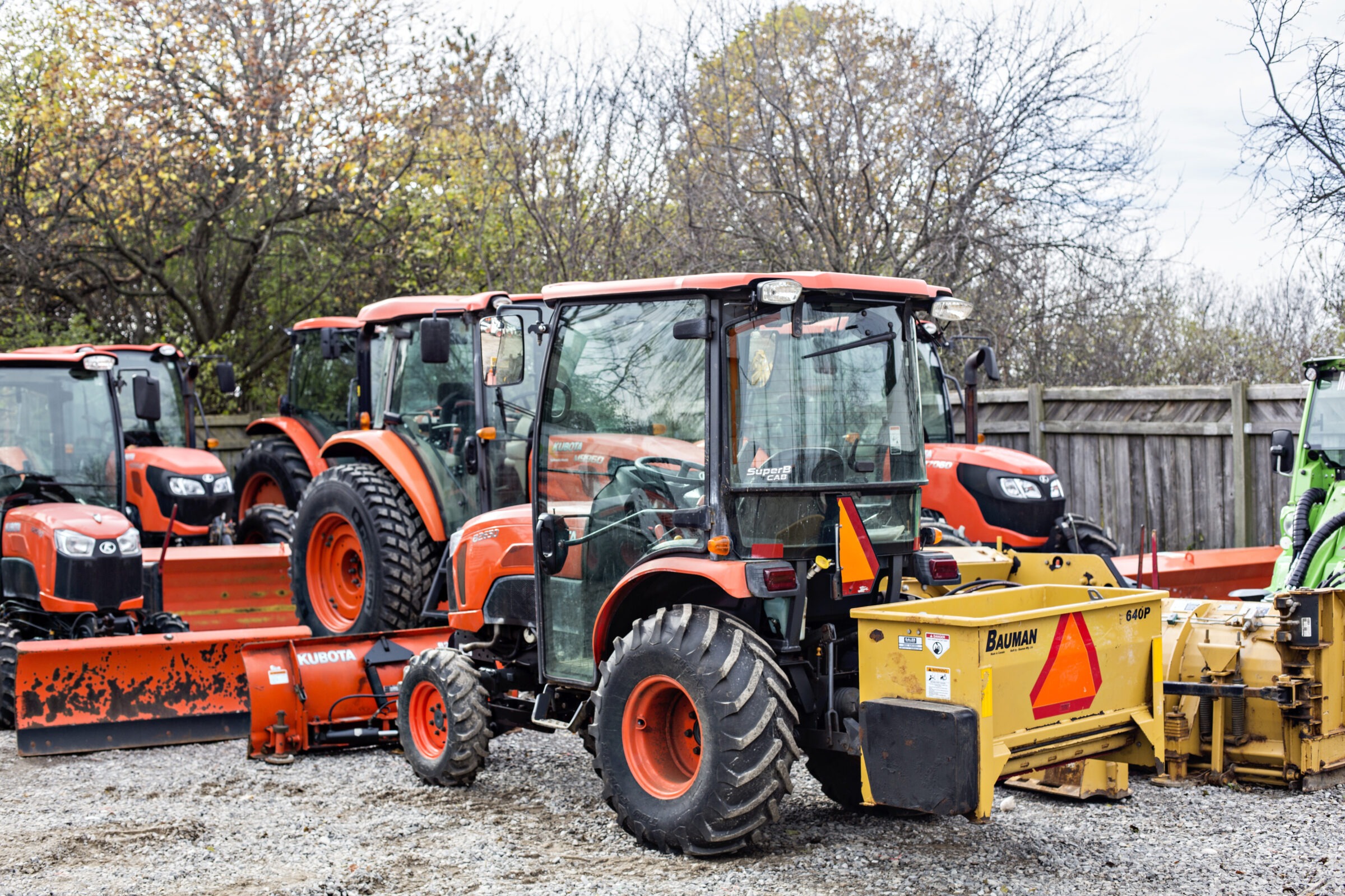 Several orange tractors, equipped with plow attachments, are parked on gravel. They are surrounded by leafless trees and a wooden fence.