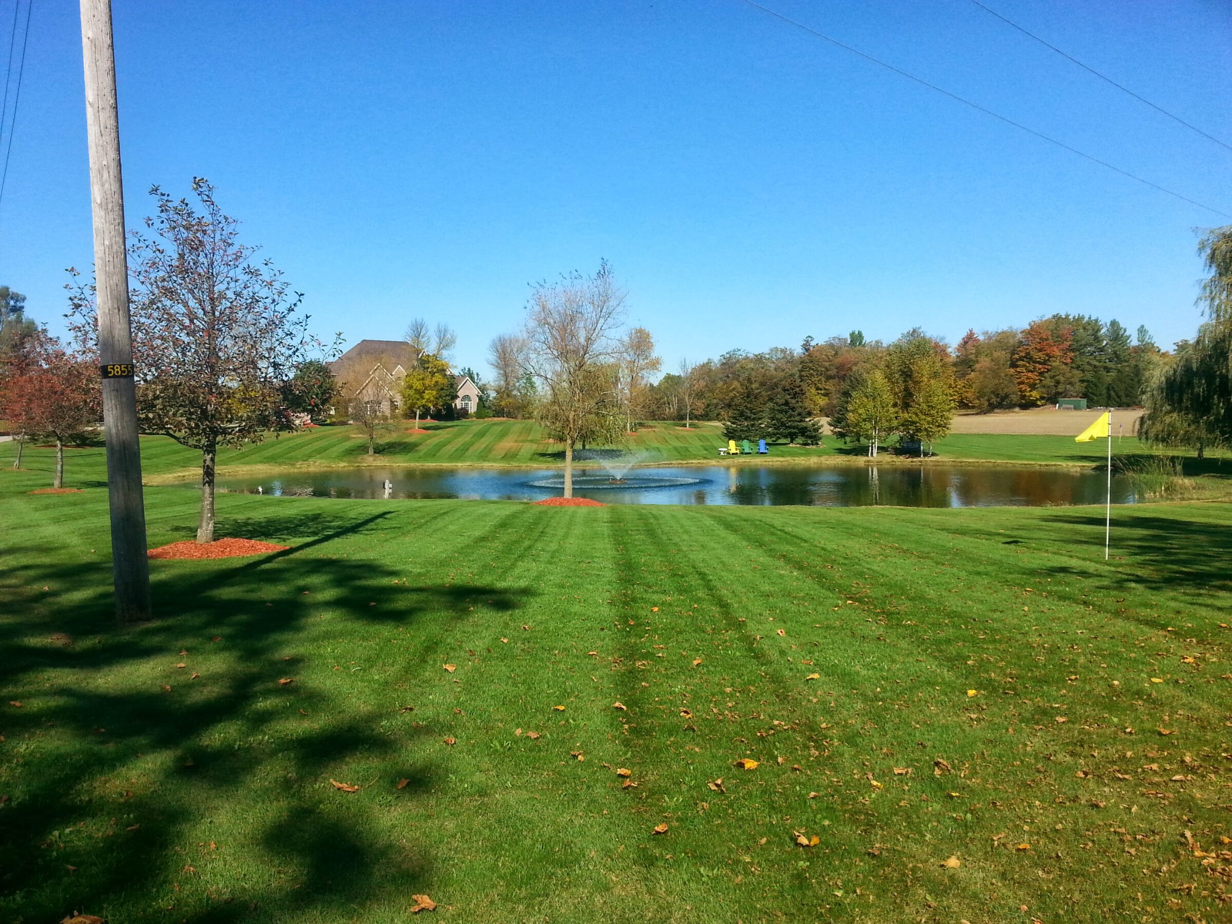 A serene landscape with manicured grass, a pond with chairs nearby, trees, and distant houses under a clear blue sky.