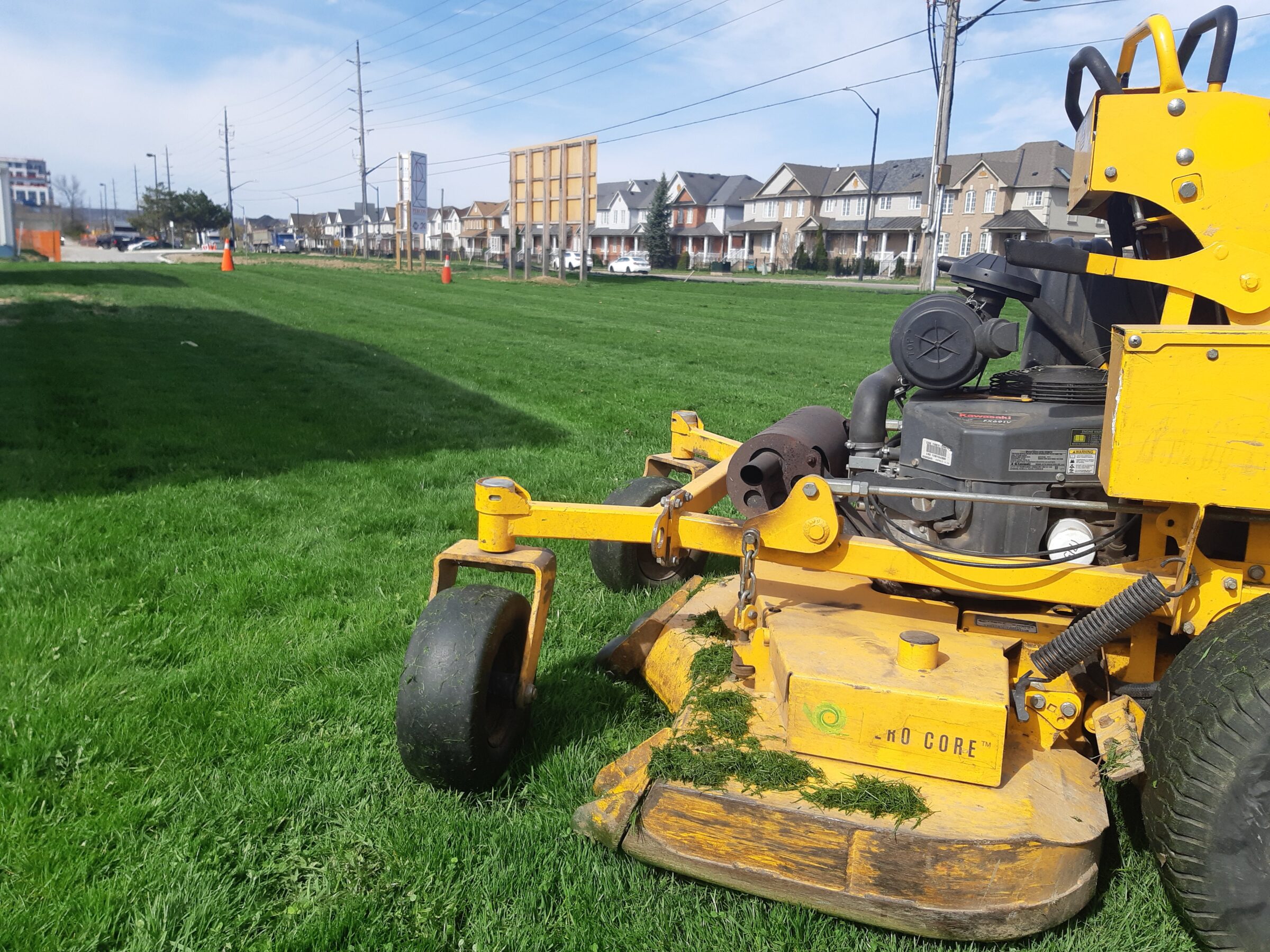 A yellow lawnmower on freshly cut grass, with suburban houses and power lines in the background under a clear blue sky.