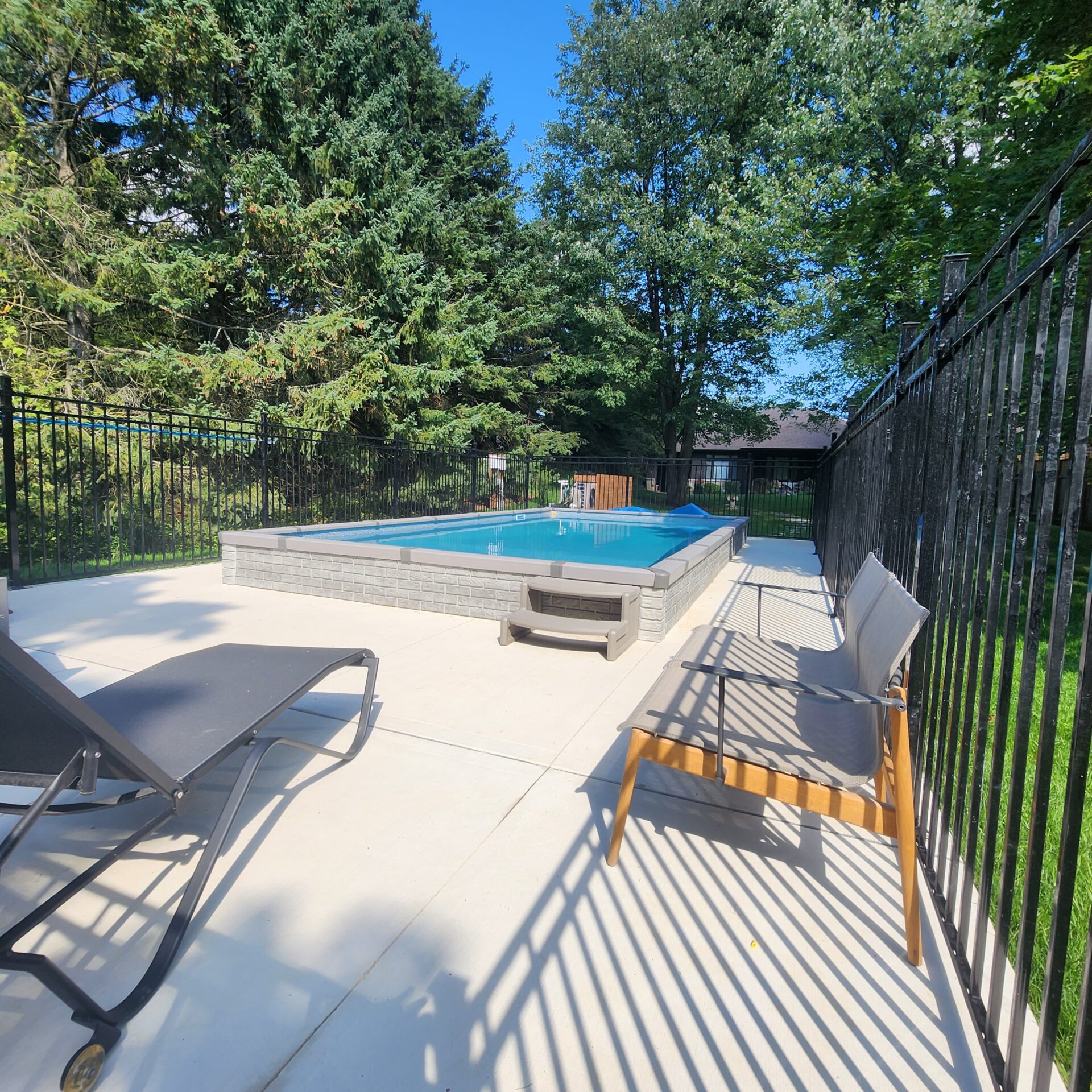 A small backyard pool surrounded by trees and chairs, enclosed by a tall black fence on a sunny day.