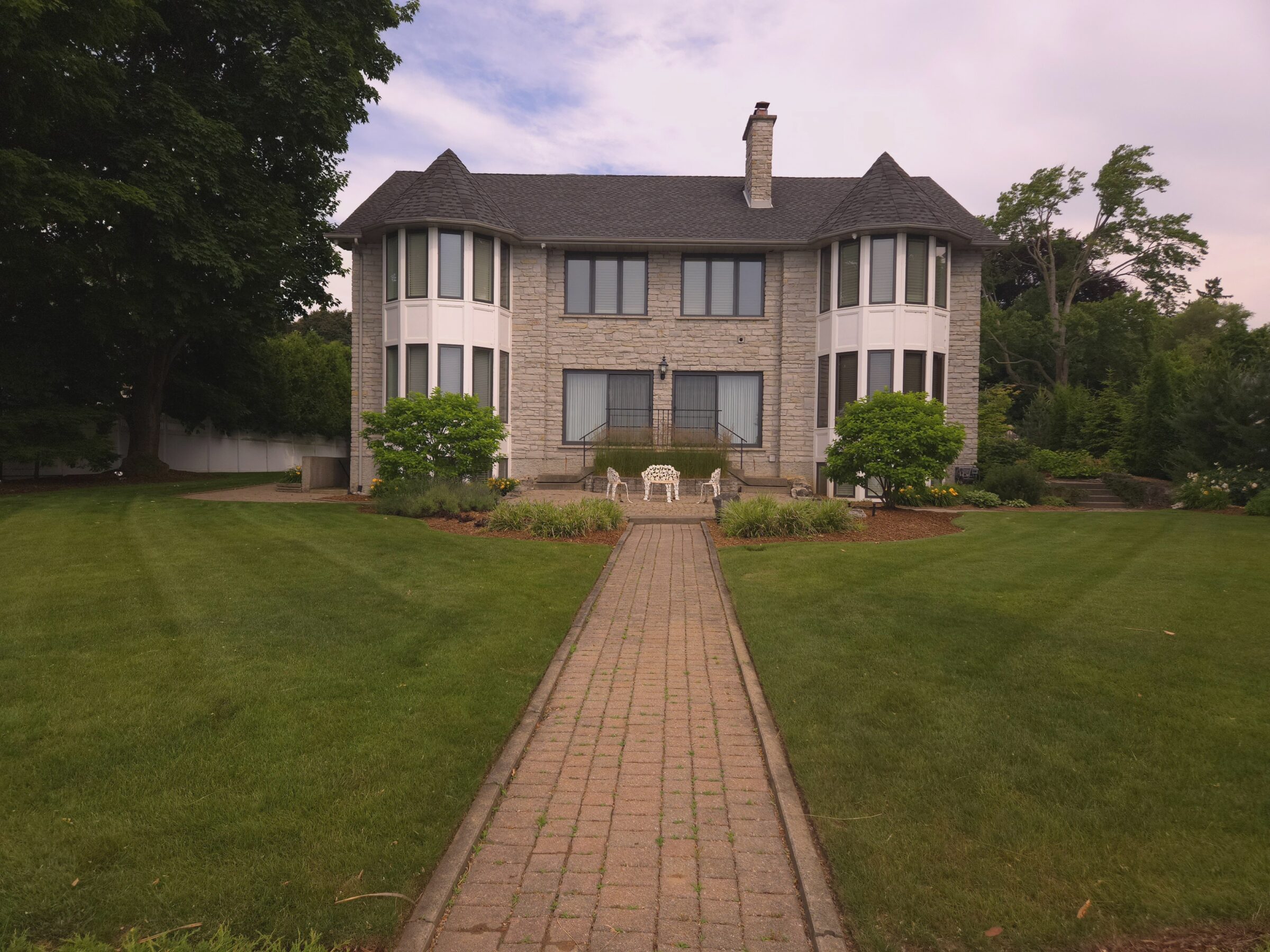 A two-story stone house with turrets, surrounded by a well-maintained lawn and trees, featuring a brick pathway leading to the entrance.