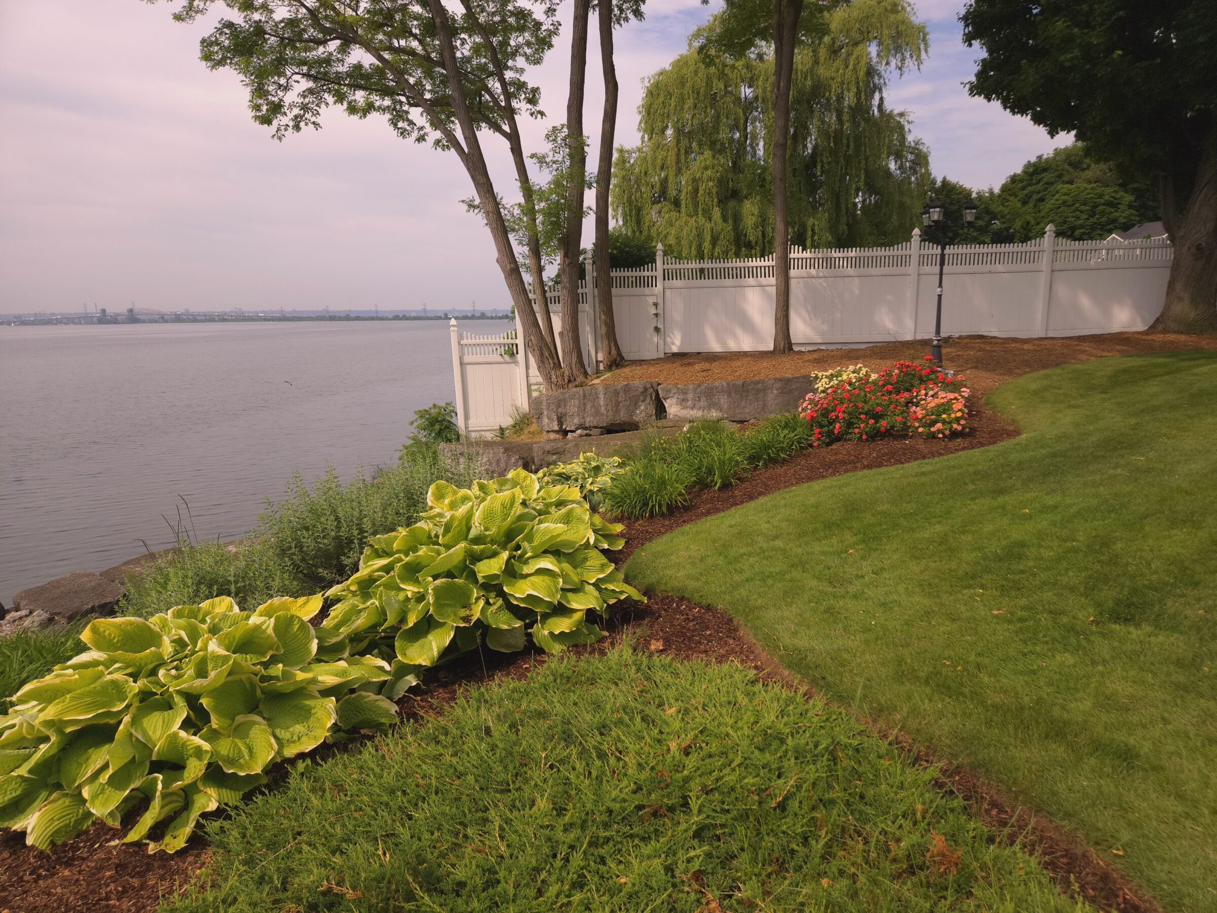 A serene garden with lush greenery overlooks a calm lake, bordered by a white fence and trees under a cloudy sky.