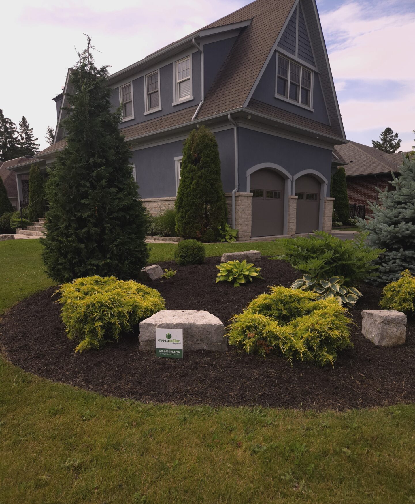 A two-story house with a steep roof, surrounded by a landscaped garden featuring shrubs and stones, set against a cloudy sky.