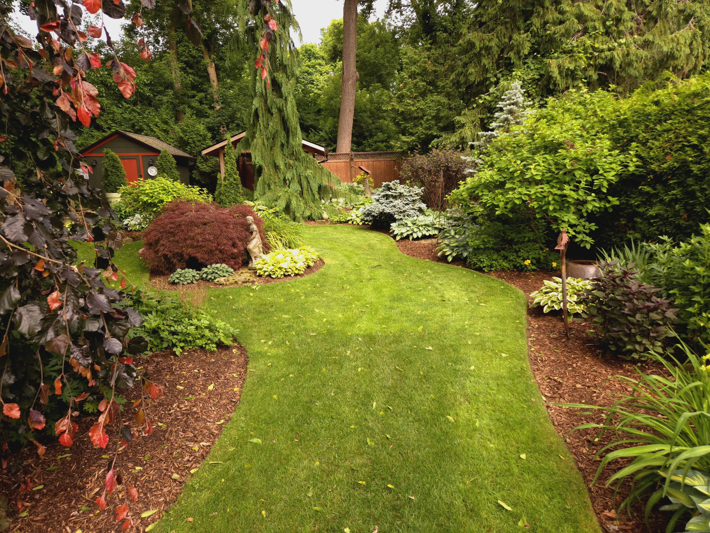 Lush garden with a winding path, vibrant foliage, and small structures surrounded by dense greenery. A serene, landscaped outdoor space.