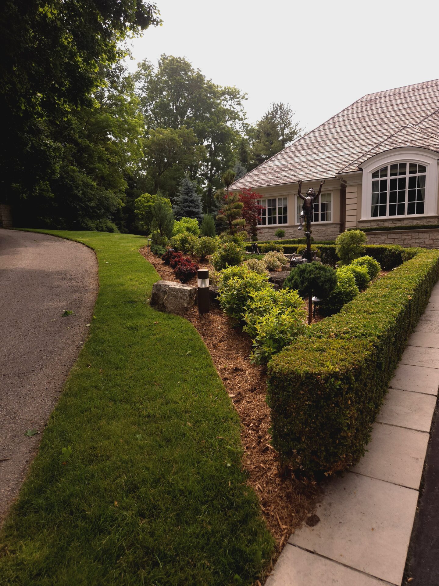 A well-maintained garden surrounds a house with a large window. A sculpture stands among the greenery, with trees lining a curved pathway.