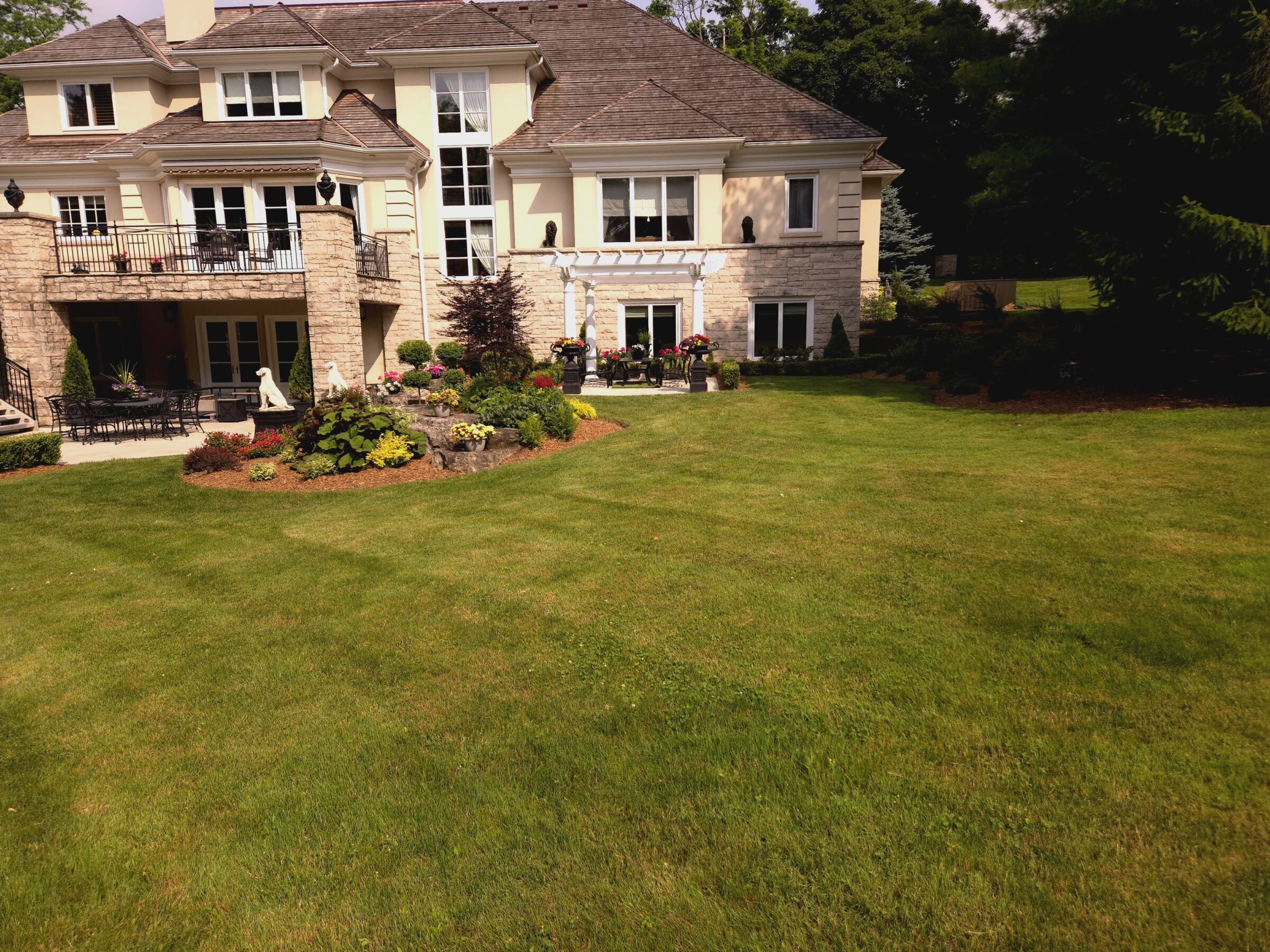 A large, elegant house with a well-kept garden, patio, and outdoor furniture, surrounded by lush greenery and vibrant flowers.