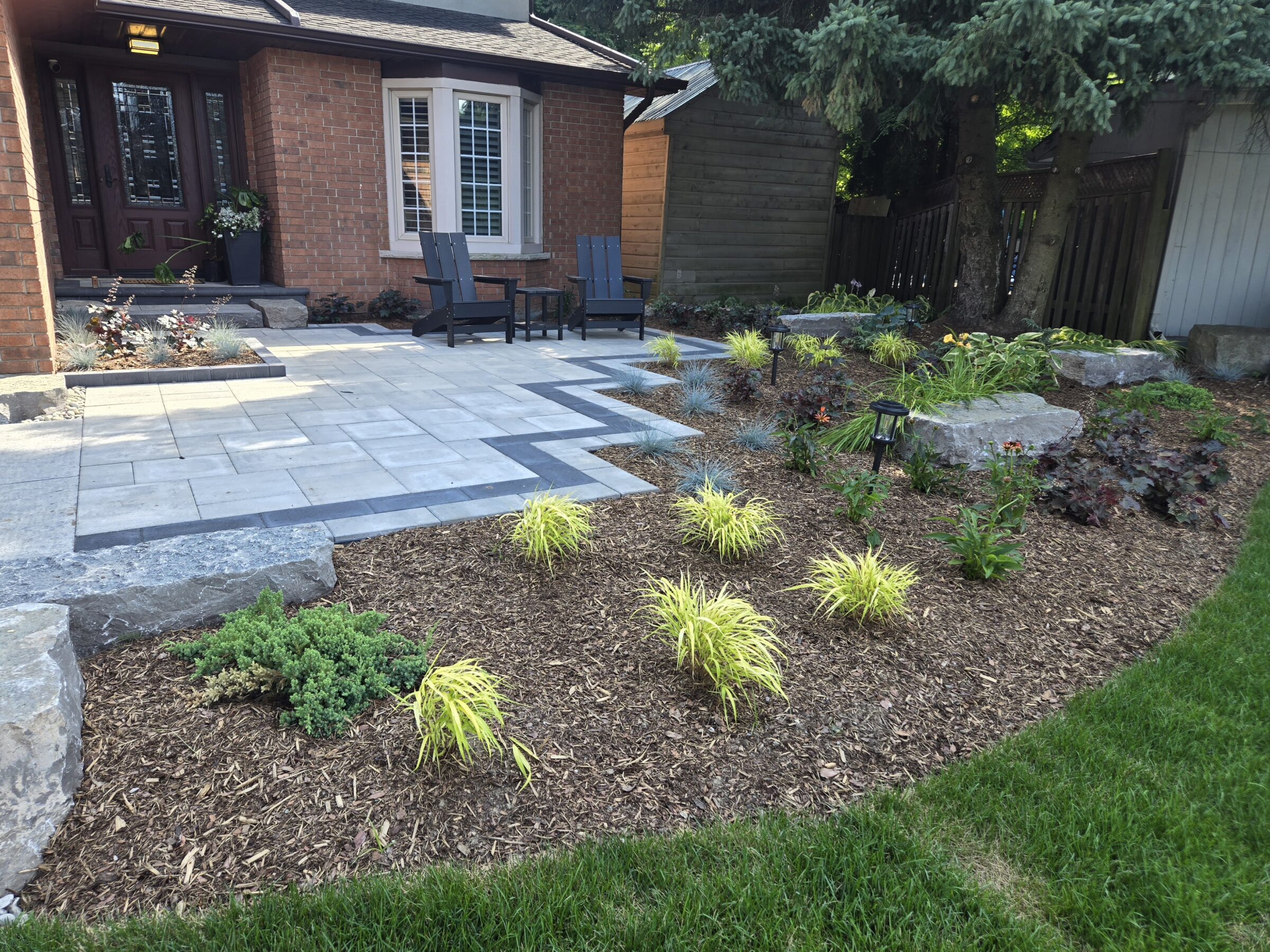 A well-maintained front yard with stone pathway, mulch garden, and plants. Two chairs are placed near the entrance of a brick house.