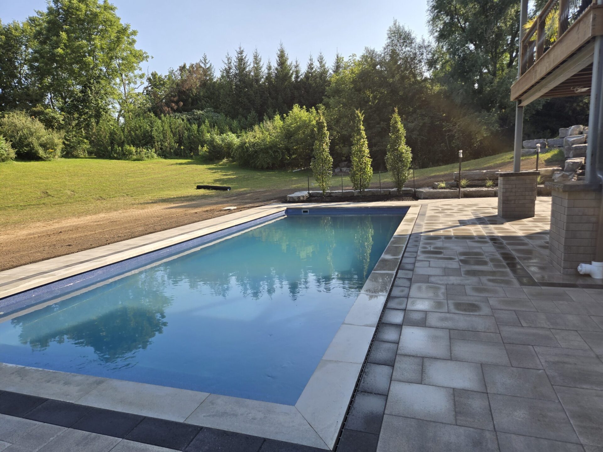 A serene backyard features a rectangular pool, surrounded by landscaped greenery and a tiled patio, with trees in the background under clear skies.