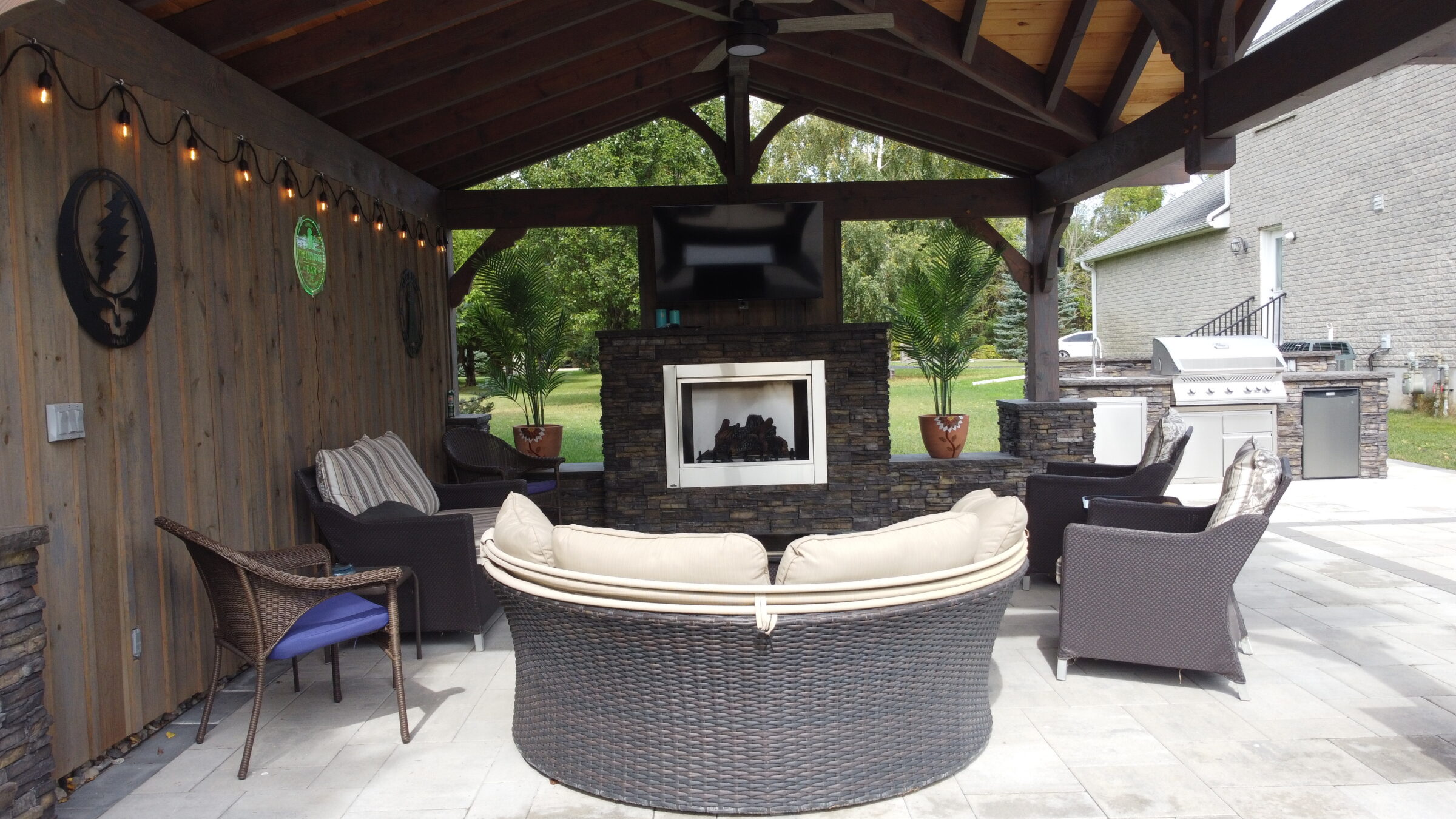 Covered outdoor seating area with fireplace, TV, and grill. Cozy chairs encircle a center table, surrounded by lush greenery and casual decor.