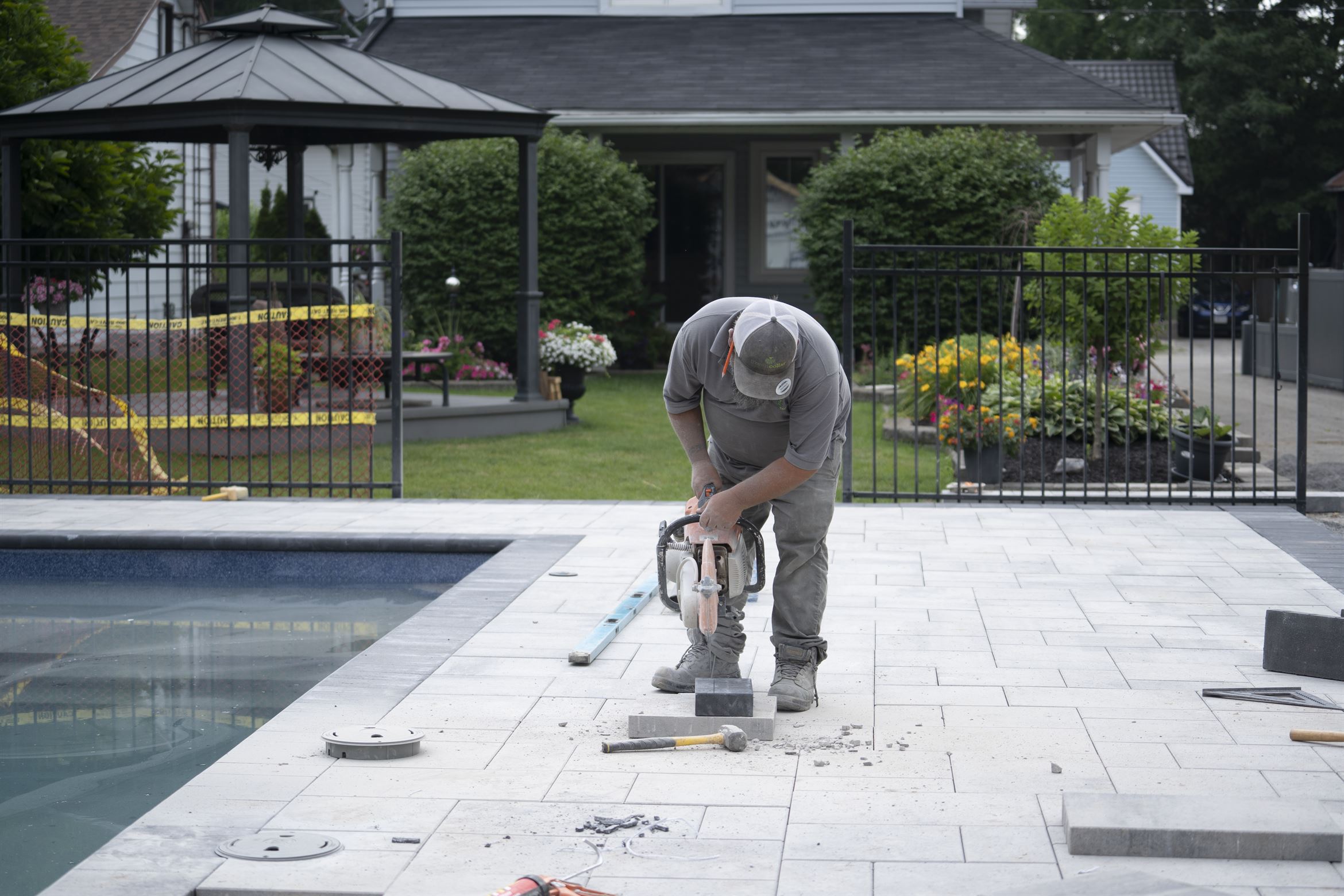 A person works with a power tool on paving stones by a pool, in a backyard with a gazebo and garden.