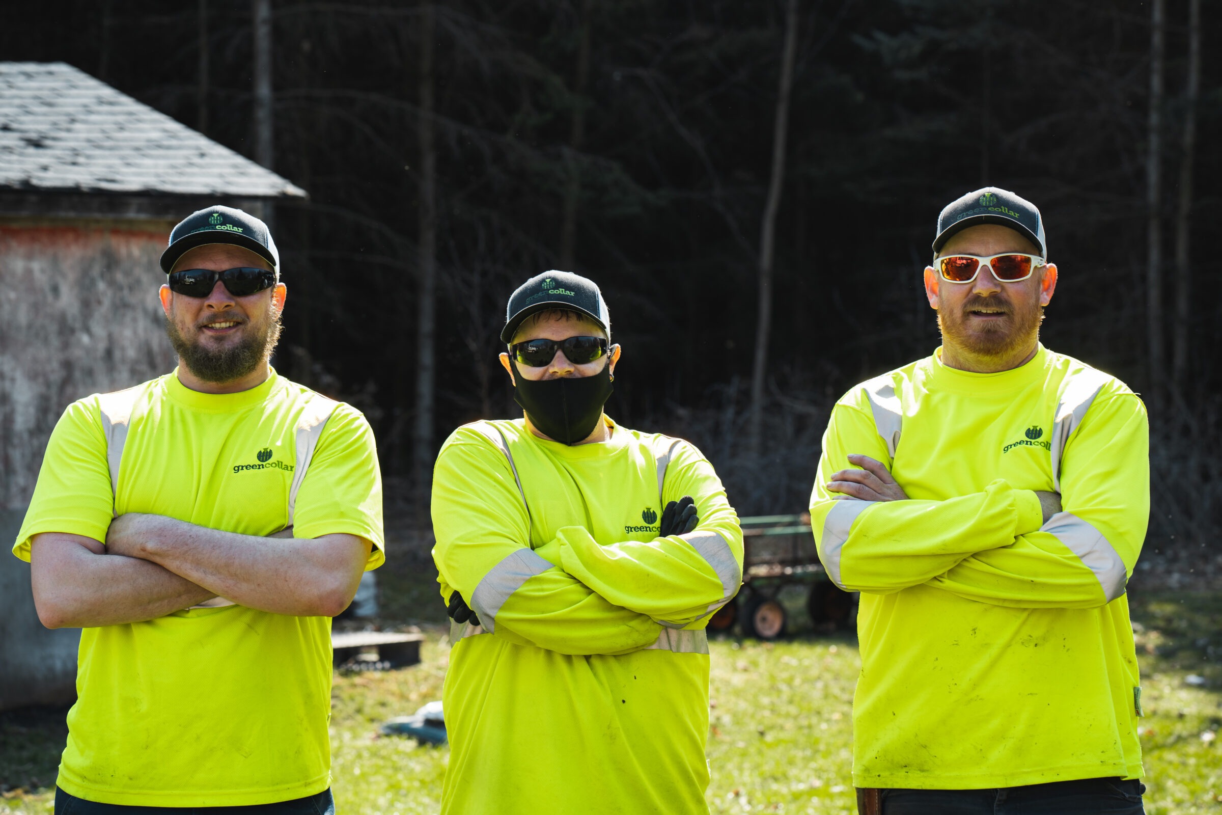 Three people in bright yellow shirts and sunglasses stand outside, arms crossed, near a small building. Forest background, no recognizable landmarks.