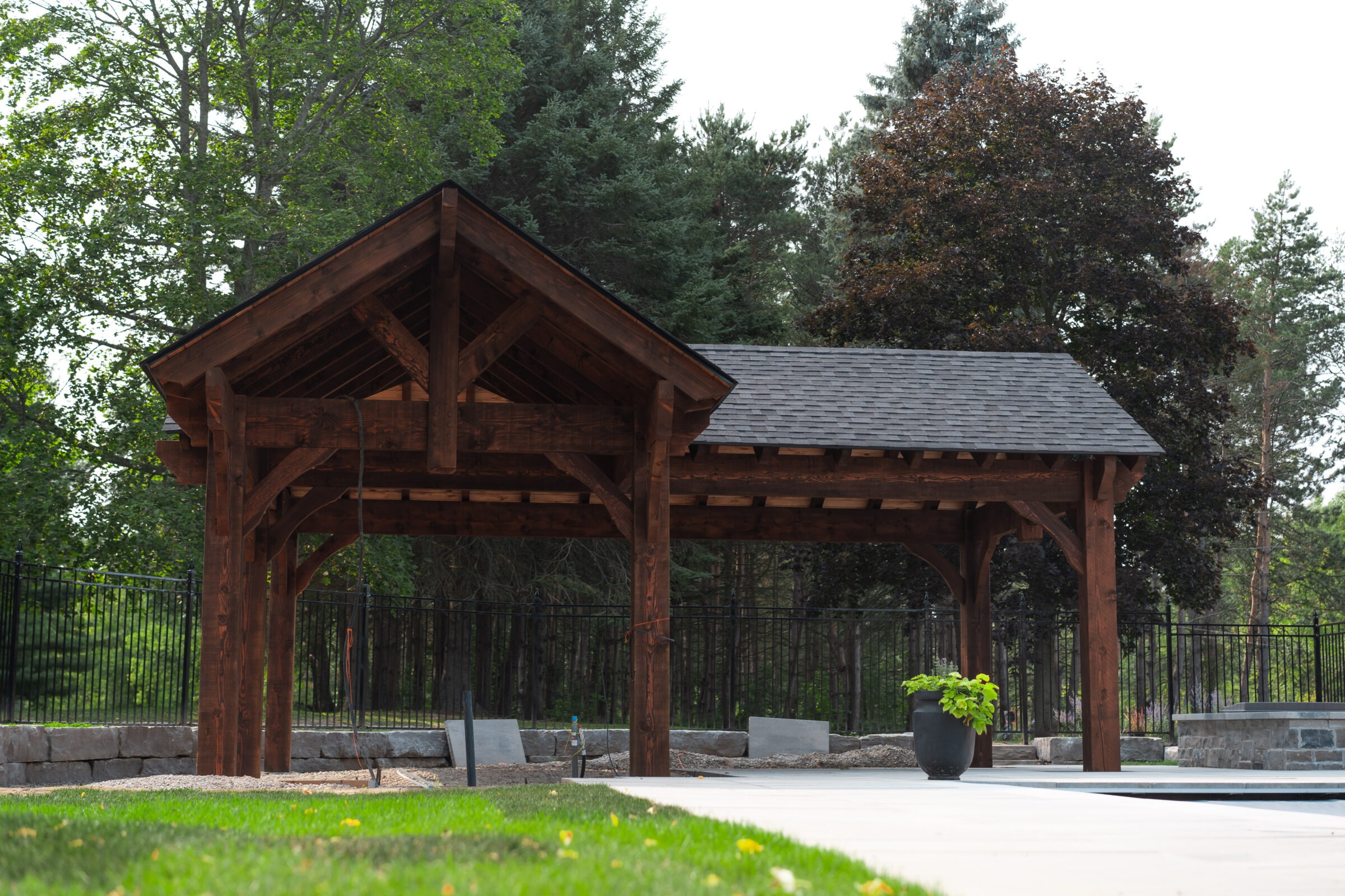 A wooden pavilion with a sloped roof stands in a landscaped garden surrounded by trees and a black metal fence.