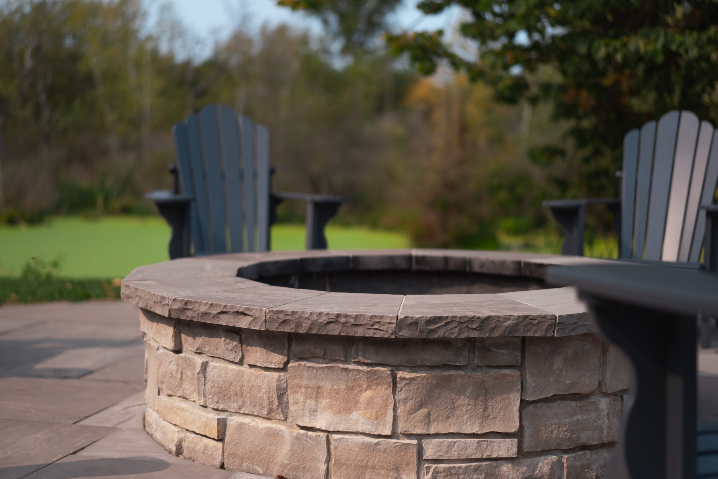 Stone fire pit with Adirondack chairs, surrounded by lush greenery and trees, creating a serene outdoor setting suitable for relaxation and social gatherings.