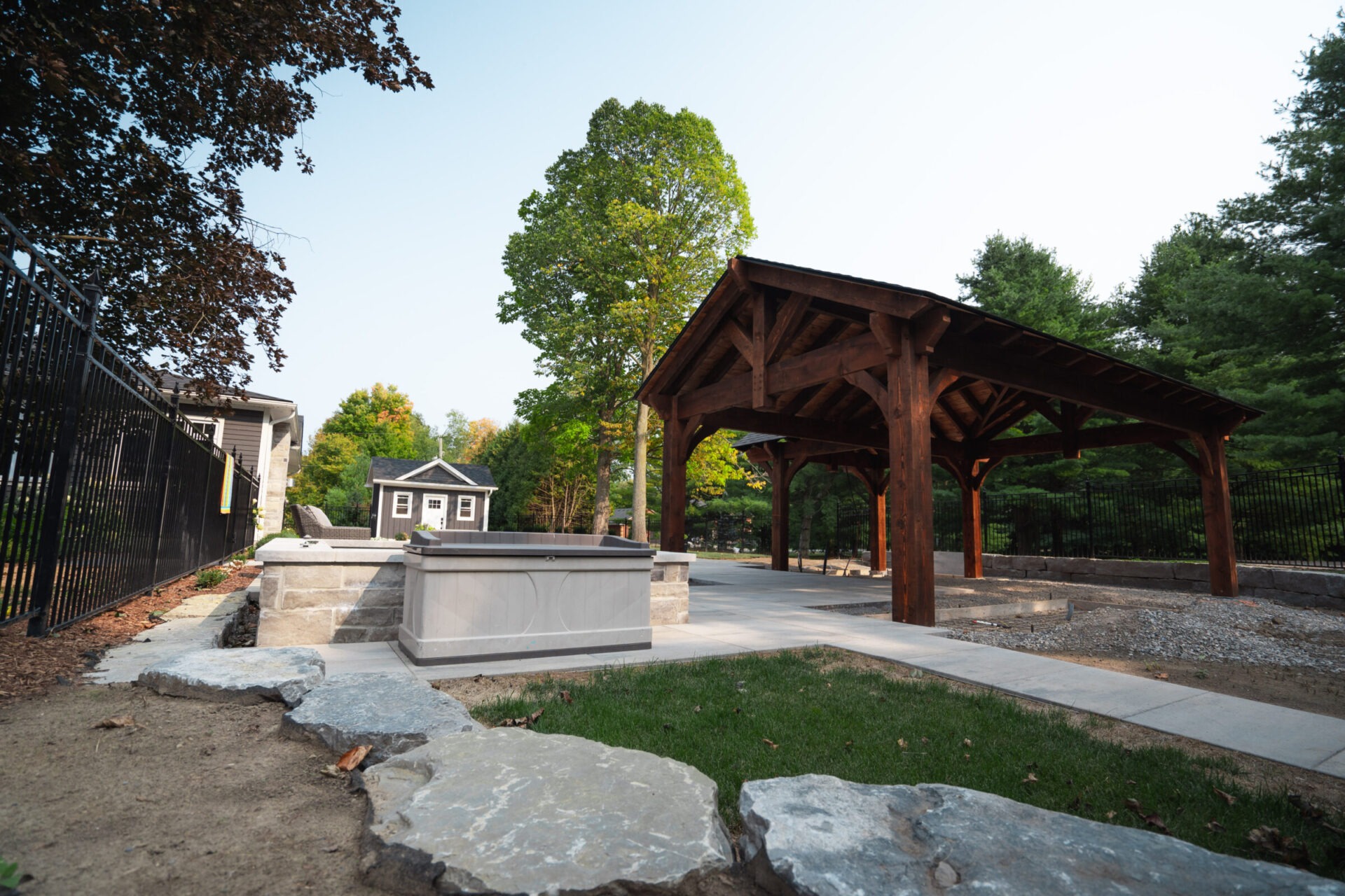 A wooden pavilion stands in a landscaped garden with stone pathways, surrounded by trees and a fence, under a clear sky.