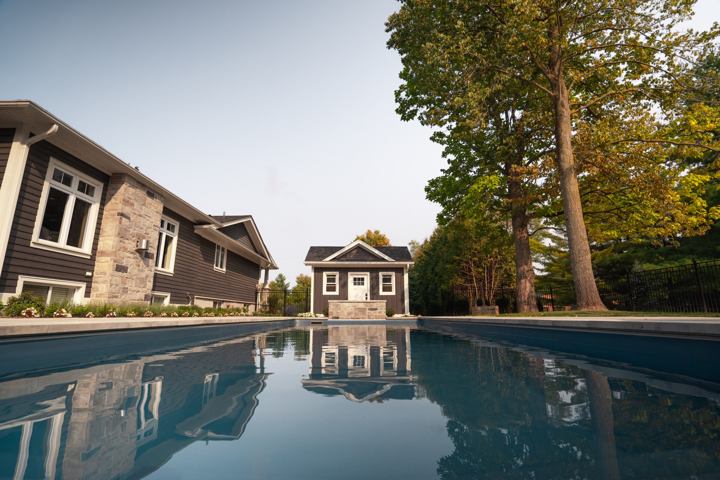 Backyard scene featuring a modern house, swimming pool, and small pool house, surrounded by lush trees under a clear sky.