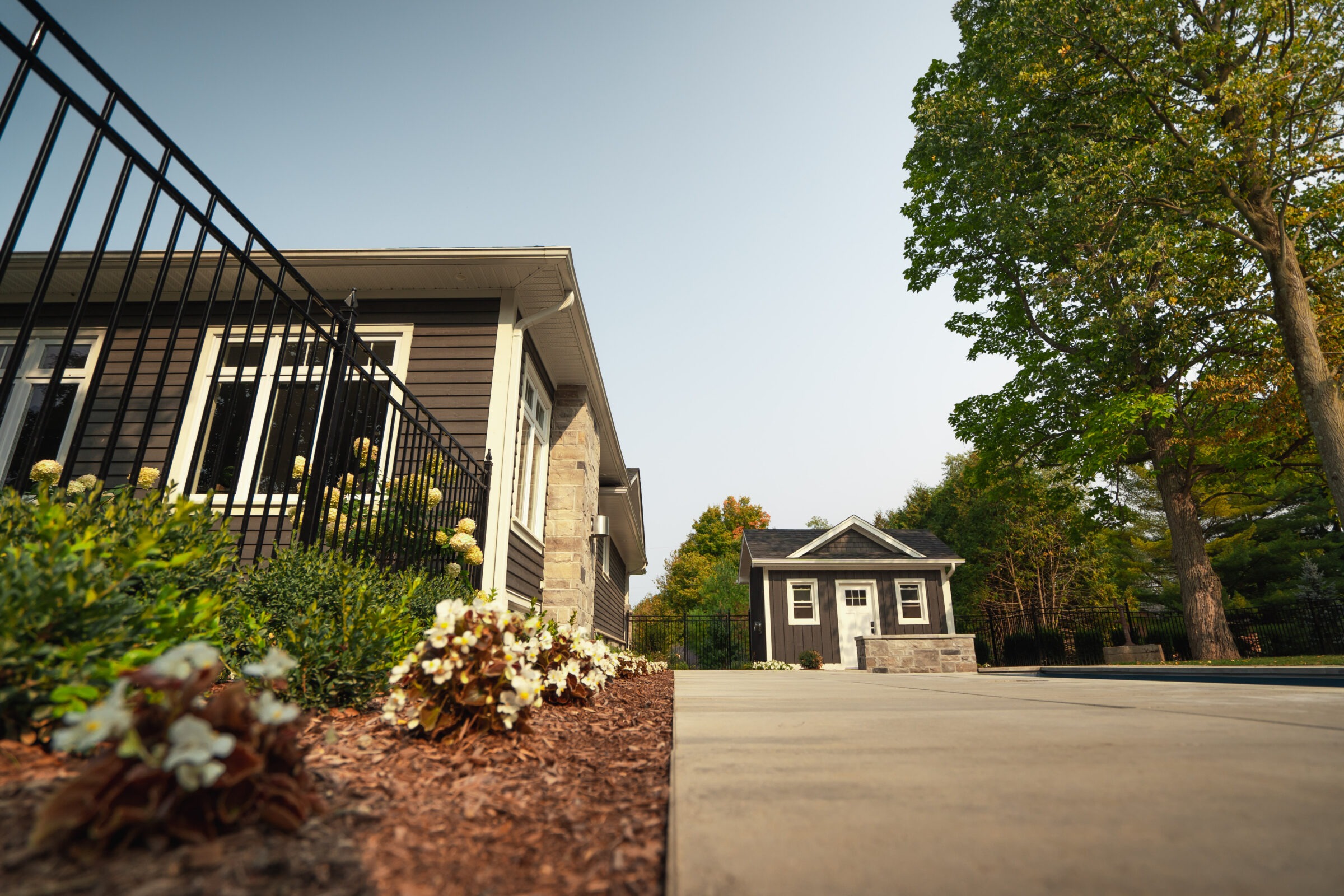 A modern house with manicured landscaping, featuring shrubs and flowers along a black fence. A small outbuilding is visible in the background.