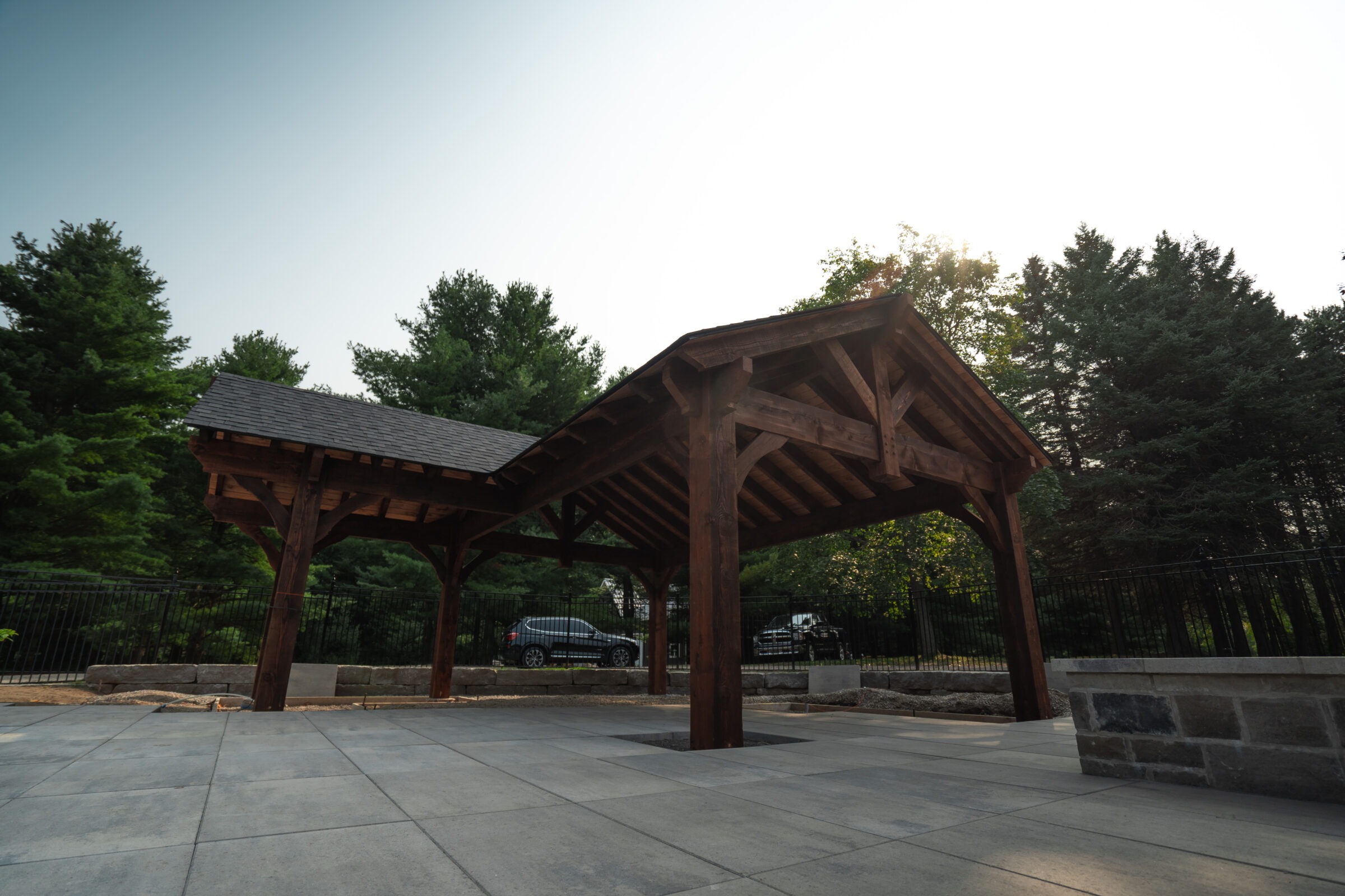 Wooden pavilion in a serene outdoor setting with surrounding trees, stone walls, and parked cars under a clear sky.