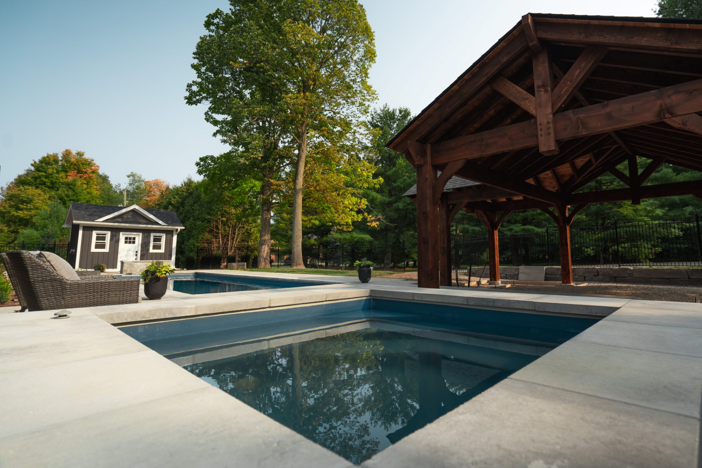 Outdoor scene with a pool, gazebo, and shed surrounded by trees, featuring a wicker chair and potted plants on the patio.