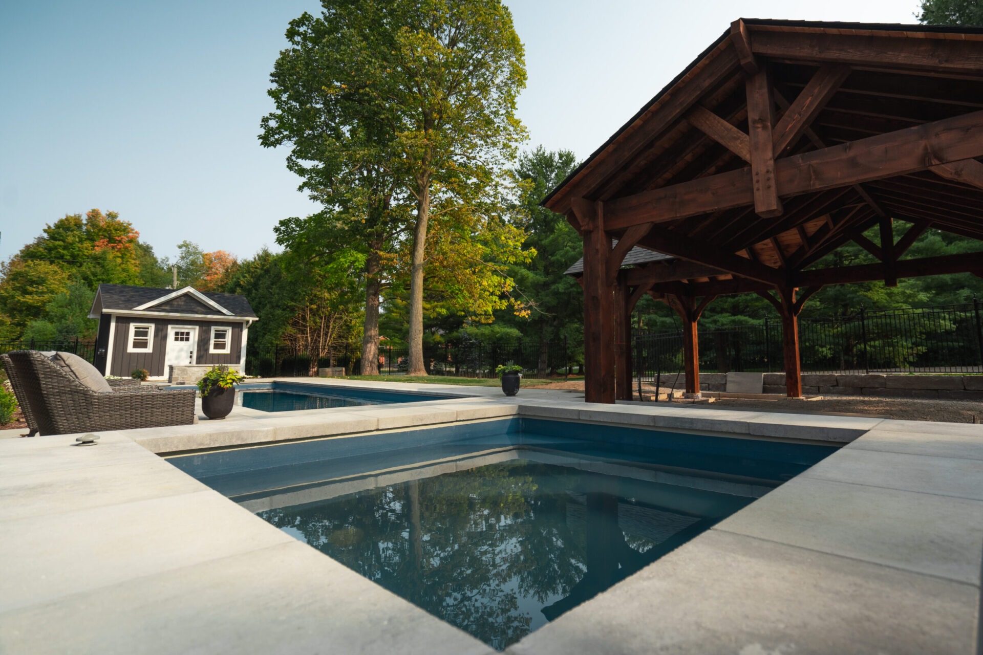 Outdoor patio with a pool, wooden pergola, and seating. A small shed is visible, surrounded by lush trees and greenery.