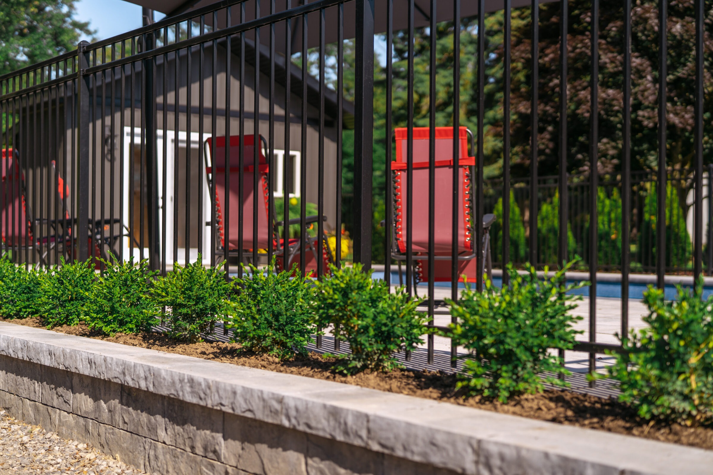 Poolside area with red lounge chairs behind a black metal fence, surrounded by lush greenery and a small building in the background.