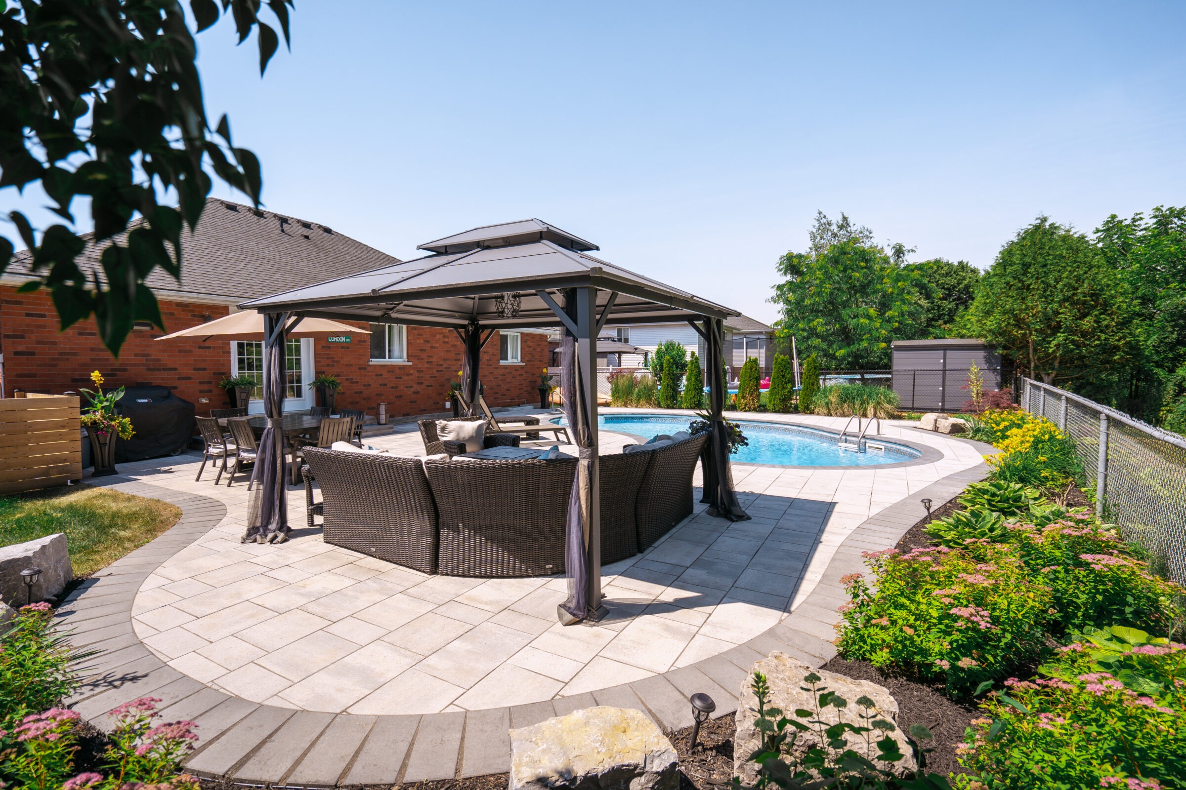 A backyard features a swimming pool, patio furniture under a gazebo, surrounded by landscaping with flowers and greenery, beside a brick house.