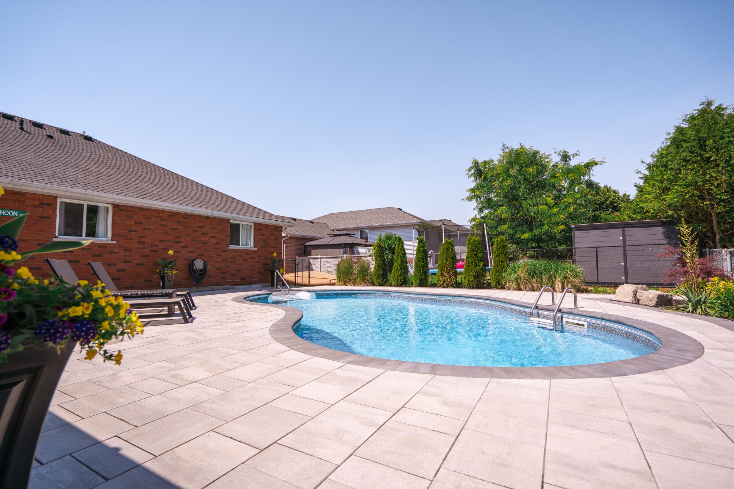 A backyard features a kidney-shaped pool, patio chairs, flowers, and a red brick house, surrounded by trees and a clear blue sky.