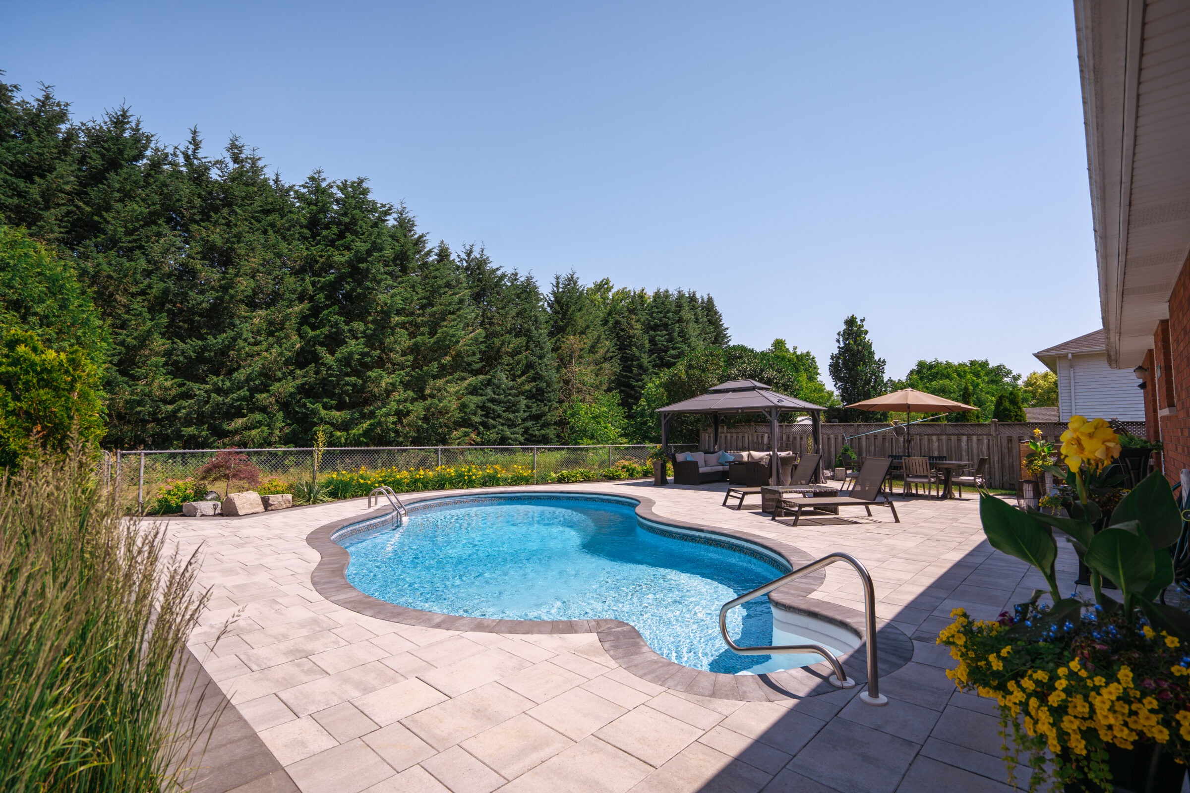 A backyard with a kidney-shaped pool, patio furniture, gazebo, and blooming flowers against lush greenery and clear blue sky.