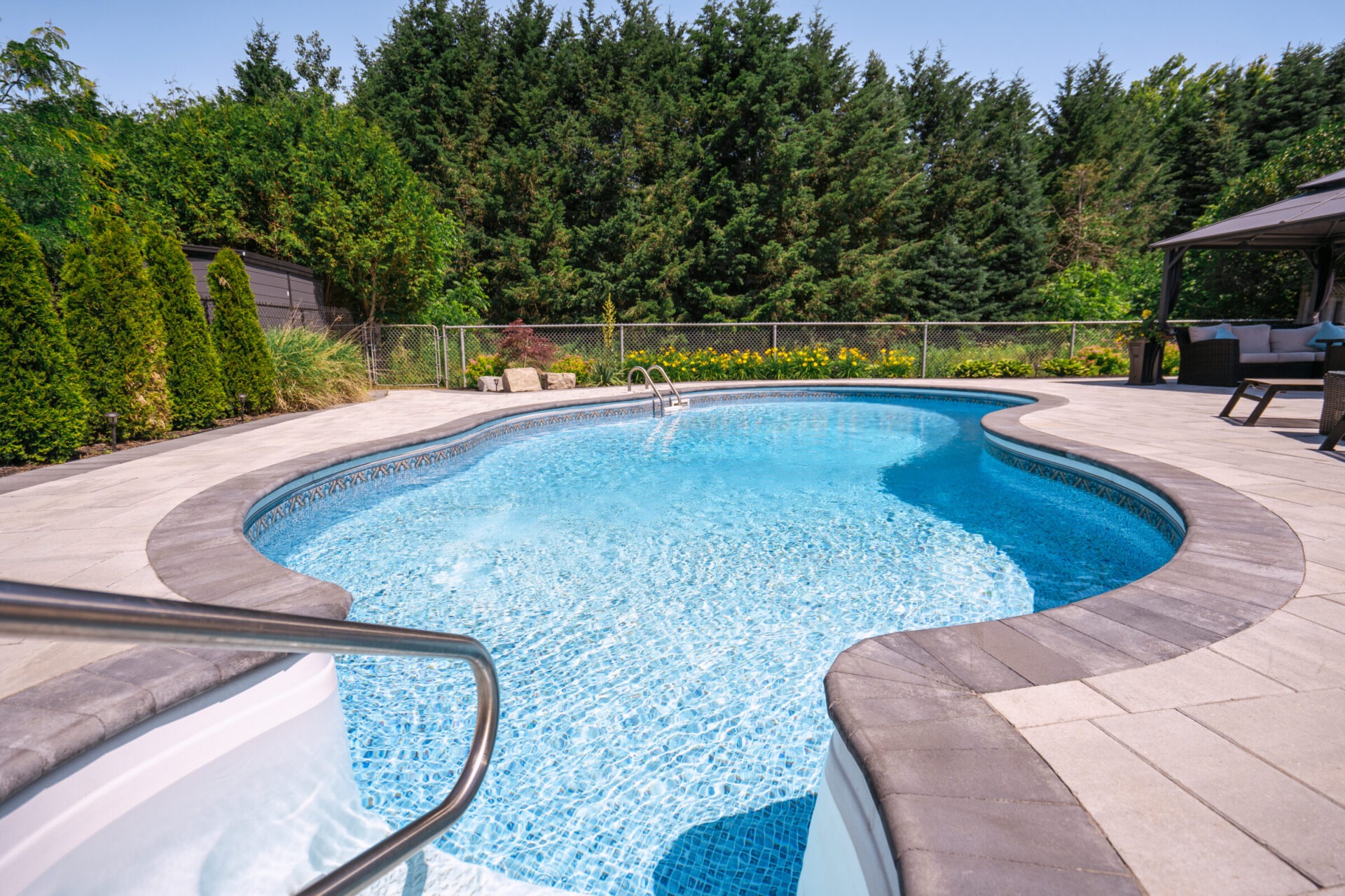 A serene backyard features a kidney-shaped swimming pool surrounded by paving stones, lush greenery, and a cozy seating area underneath a gazebo.