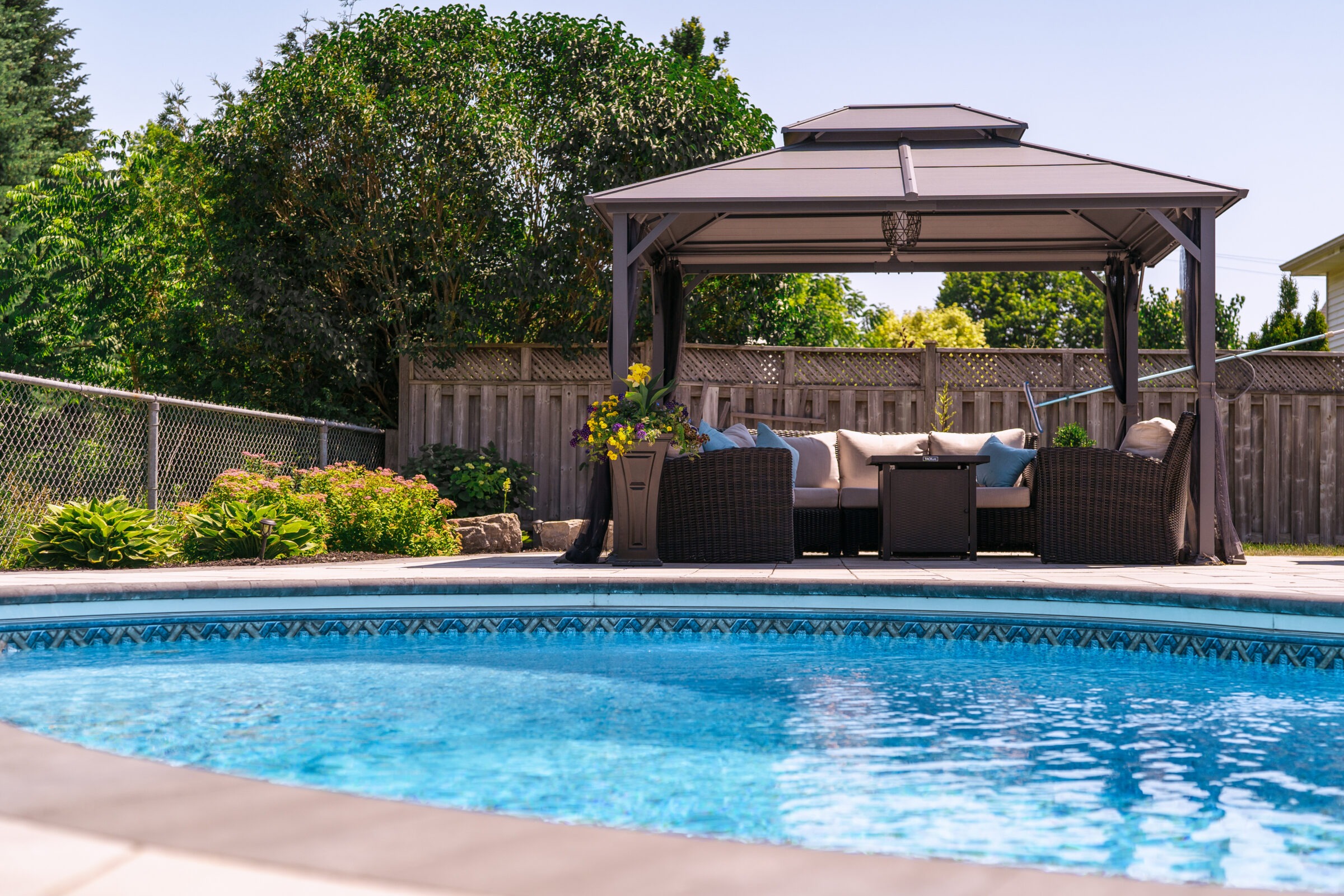 A backyard pool with clear water, surrounded by seating under a modern gazebo with vibrant flowers and lush greenery, enclosed by a wooden fence.