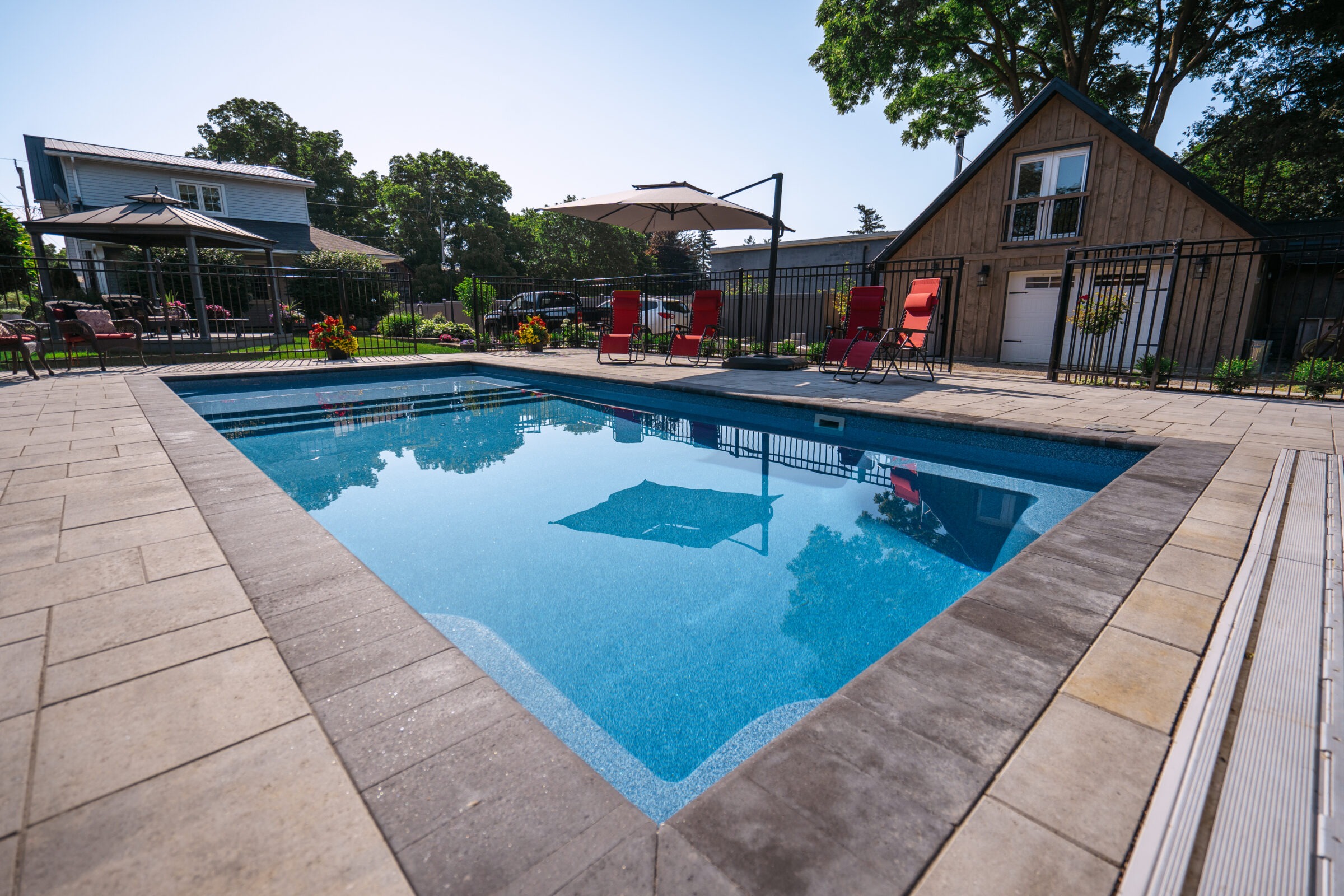 A serene backyard pool scene with red lounge chairs, umbrella, a cozy wooden house, and lush greenery under a clear blue sky.