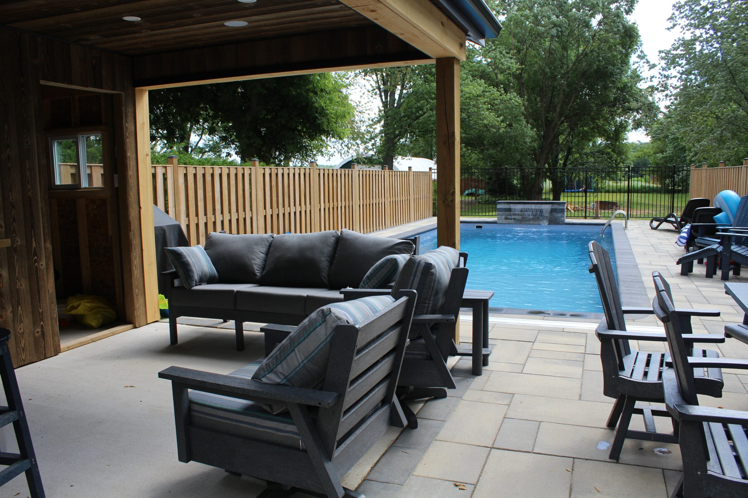 A backyard setup featuring a pool, lounge chairs, and a pergola with comfortable seating, surrounded by a wooden fence and trees.