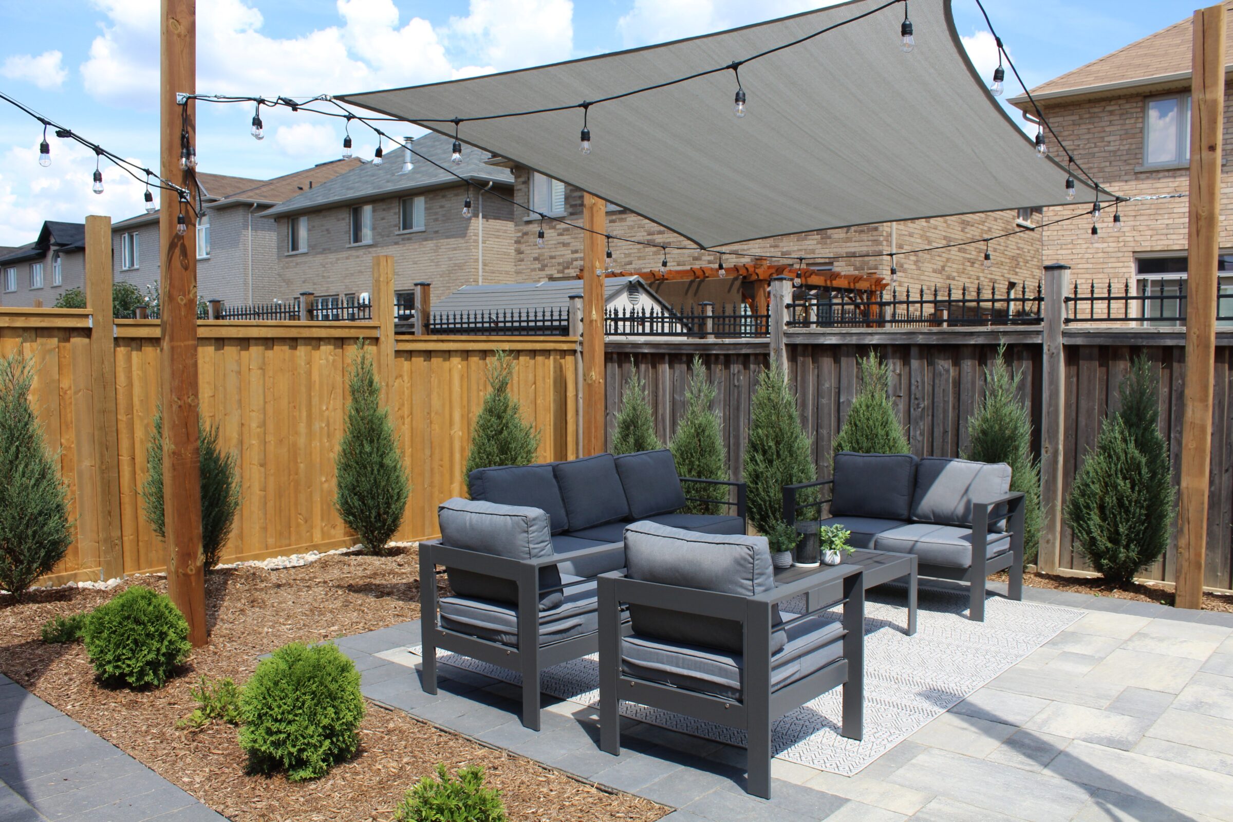 A cozy outdoor patio with gray furniture, string lights, and shade sail, surrounded by a wooden fence and brick houses.