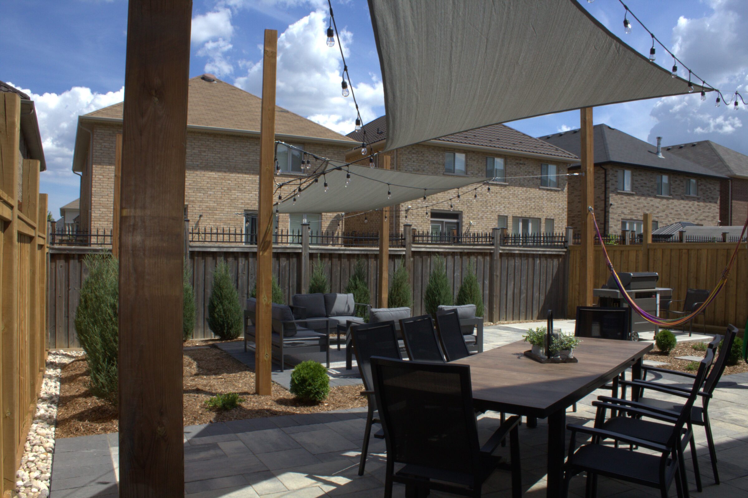 Outdoor patio with dining area, couches, and string lights. Shade sails overhead, brick houses in the background, fenced yard, landscaped with shrubs.