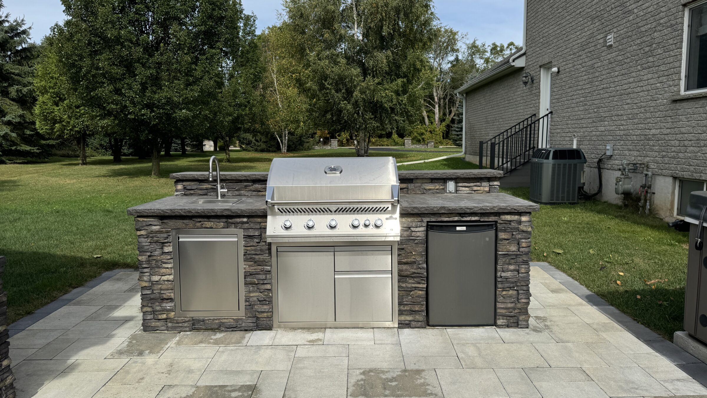 Outdoor stone kitchen with grill, fridge, and sink on a patio, surrounded by grass and trees, adjacent to a brick house.