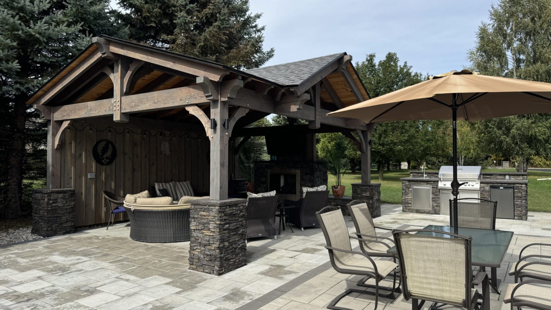 A wooden gazebo with patio furniture and barbecue area, surrounded by trees, under a clear sky, creating a cozy outdoor setting.