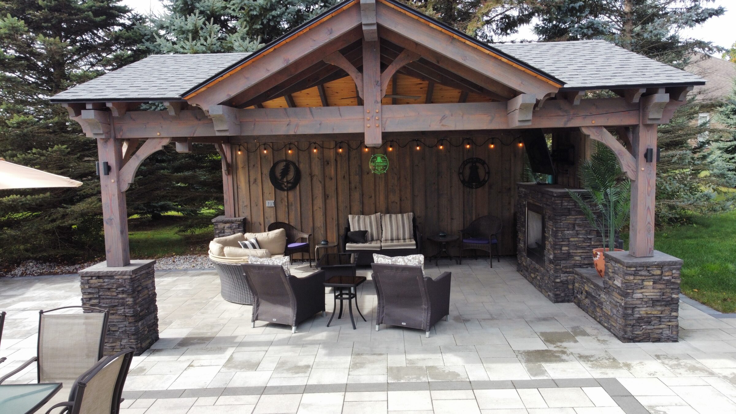 A cozy outdoor patio features a wooden gazebo, wicker furniture, and stone accents, surrounded by trees in a landscaped garden setting.