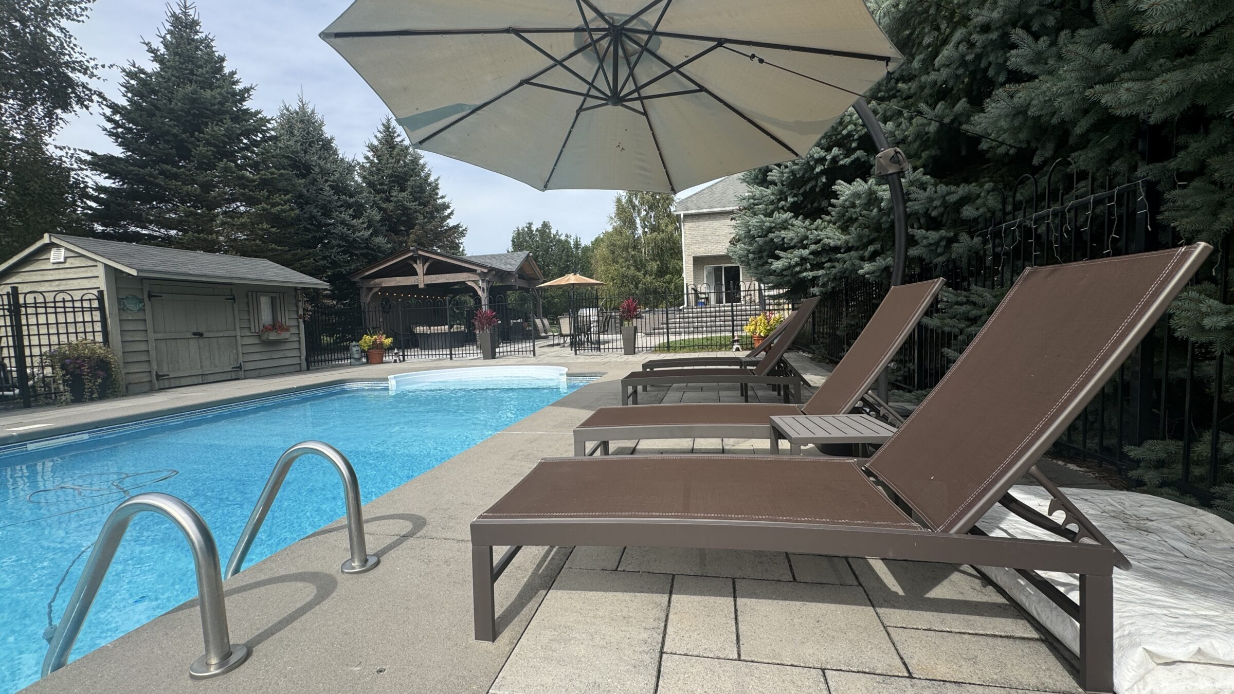 Poolside scene with lounge chairs, large umbrella, garden shed, and trees. Tranquil setting with a pavilion and decorative plants.