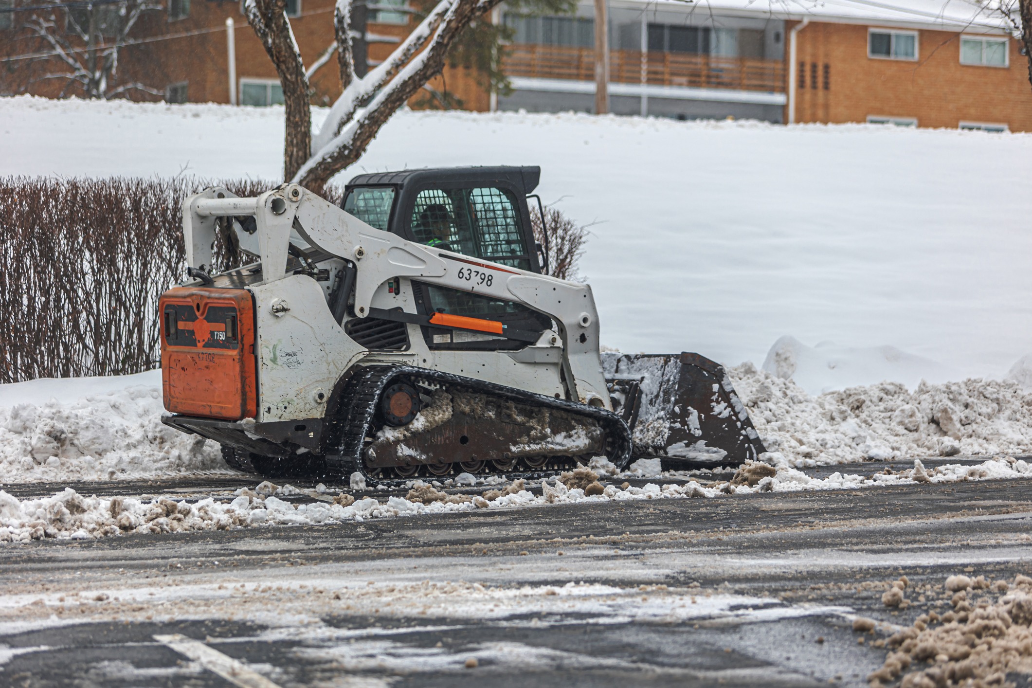 A person operates a skid-steer loader, clearing snow from a road. Snowy landscape with trees and buildings in the background.