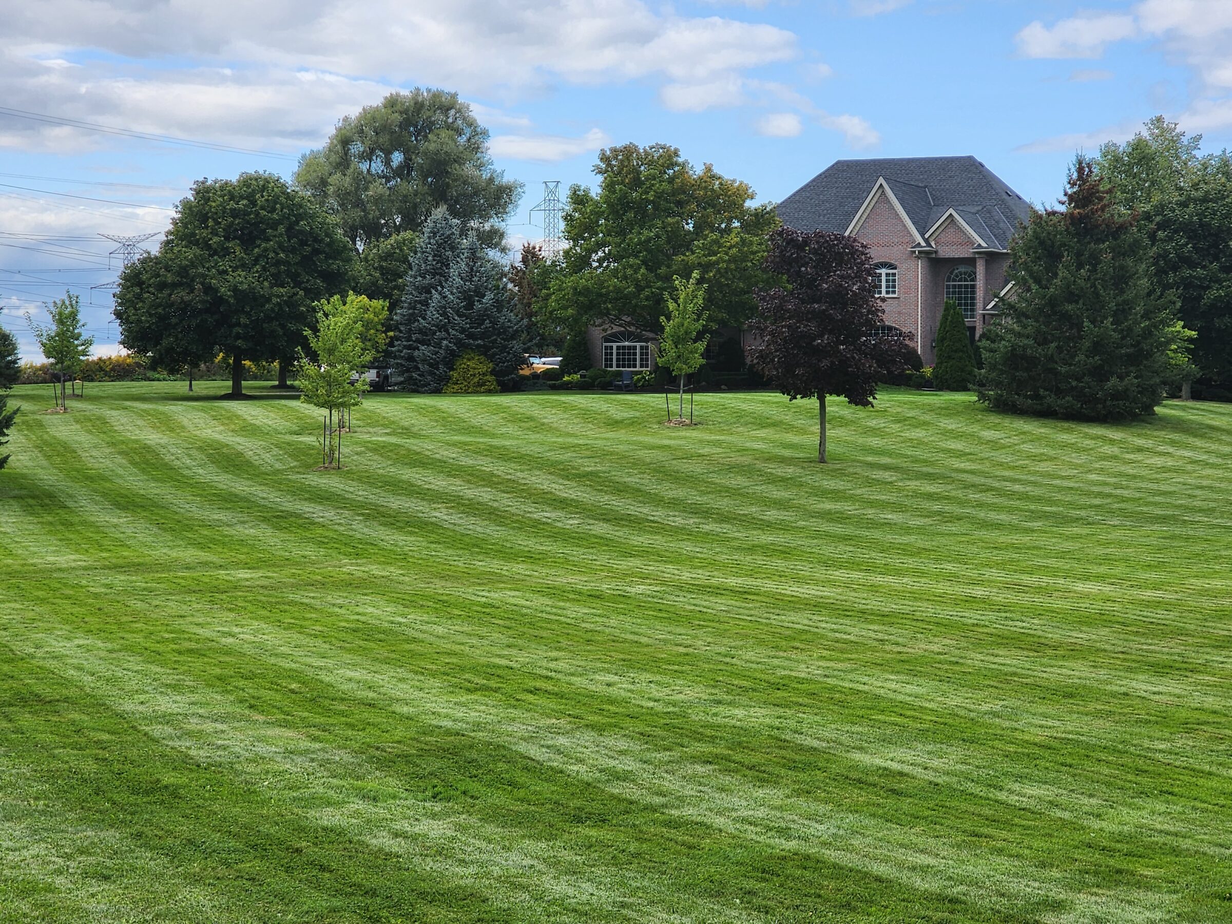 A large, well-manicured lawn with striped grass patterns, surrounded by trees, leads to a brick house under a partly cloudy sky.