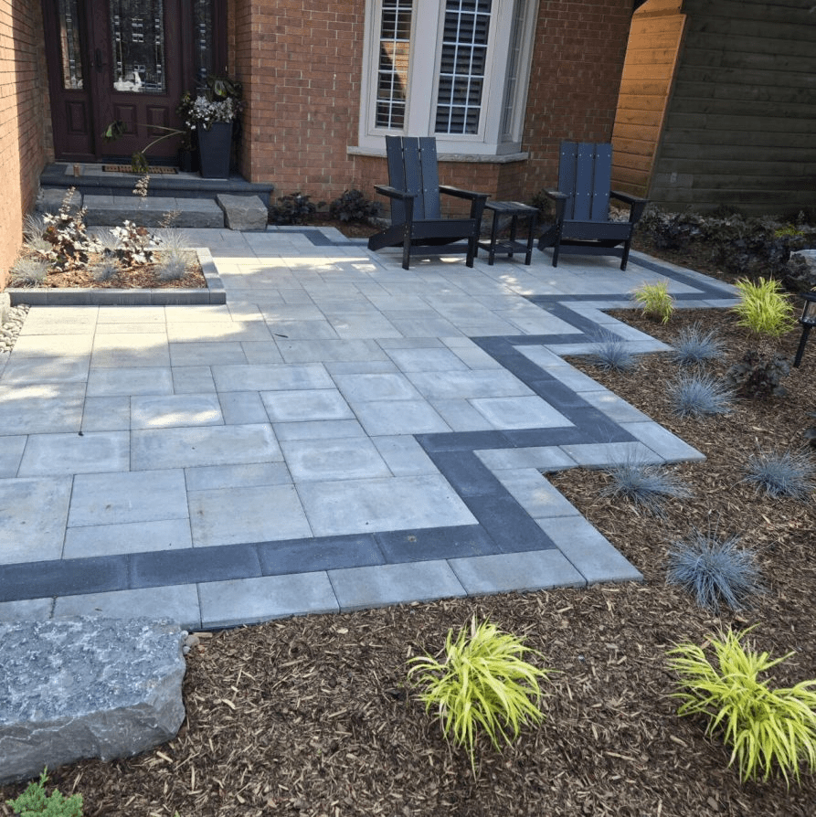 A patio area outside a brick home, featuring light grey stone pavers with a decorative dark grey zig-zag border. Two black Adirondack chairs sit on the patio, adjacent to garden beds filled with mulch and ornamental grasses.