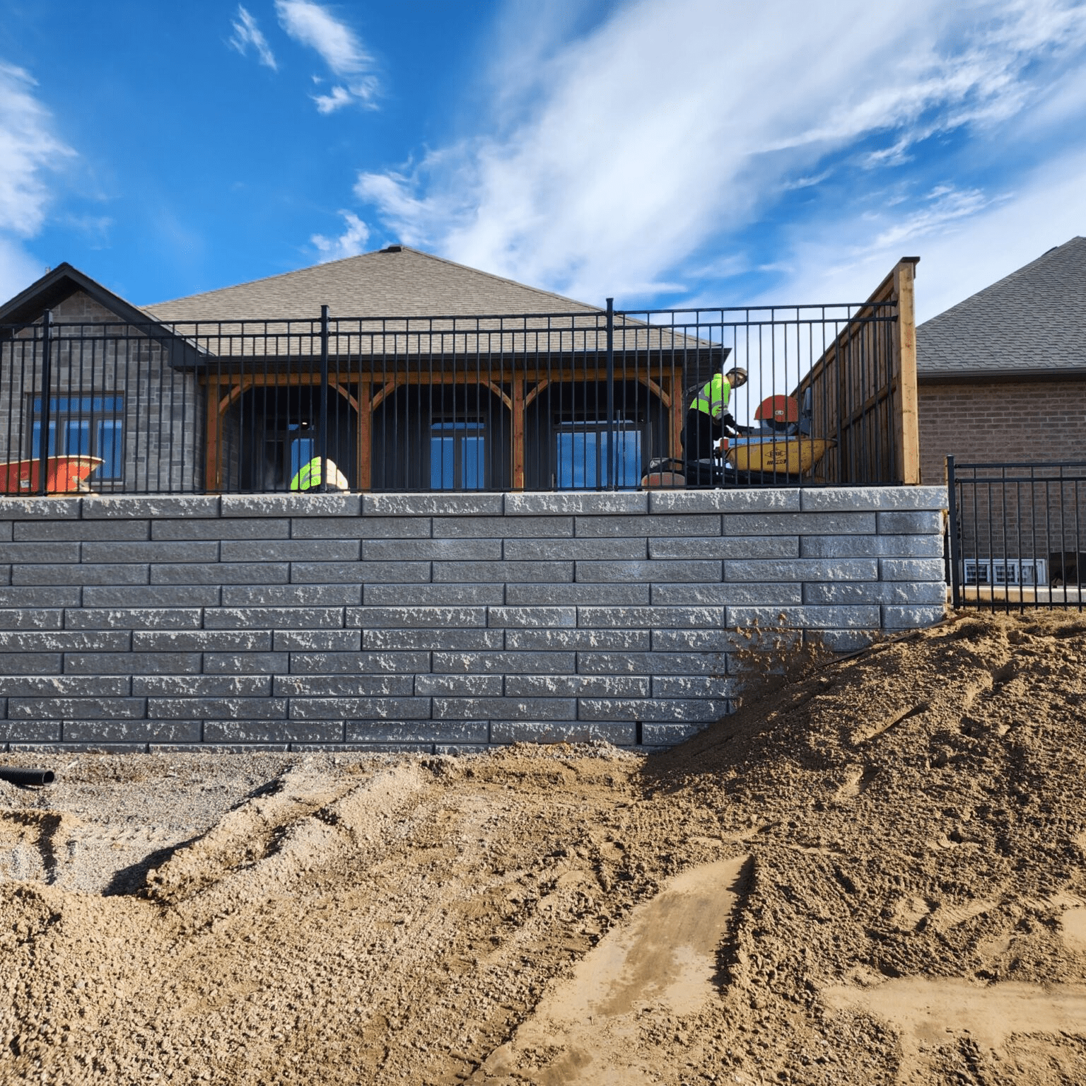 A construction site showing the build of a retaining wall made of stacked grey stone blocks. A worker in a high-visibility jacket is operating equipment behind a black metal fence on a raised deck.