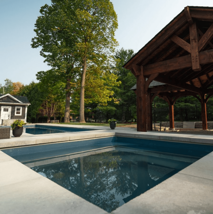 A modern backyard scene featuring a square pool or hot tub in the foreground with stone coping. In the background, there is a large wooden pavilion and a small outbuilding surrounded by mature trees.