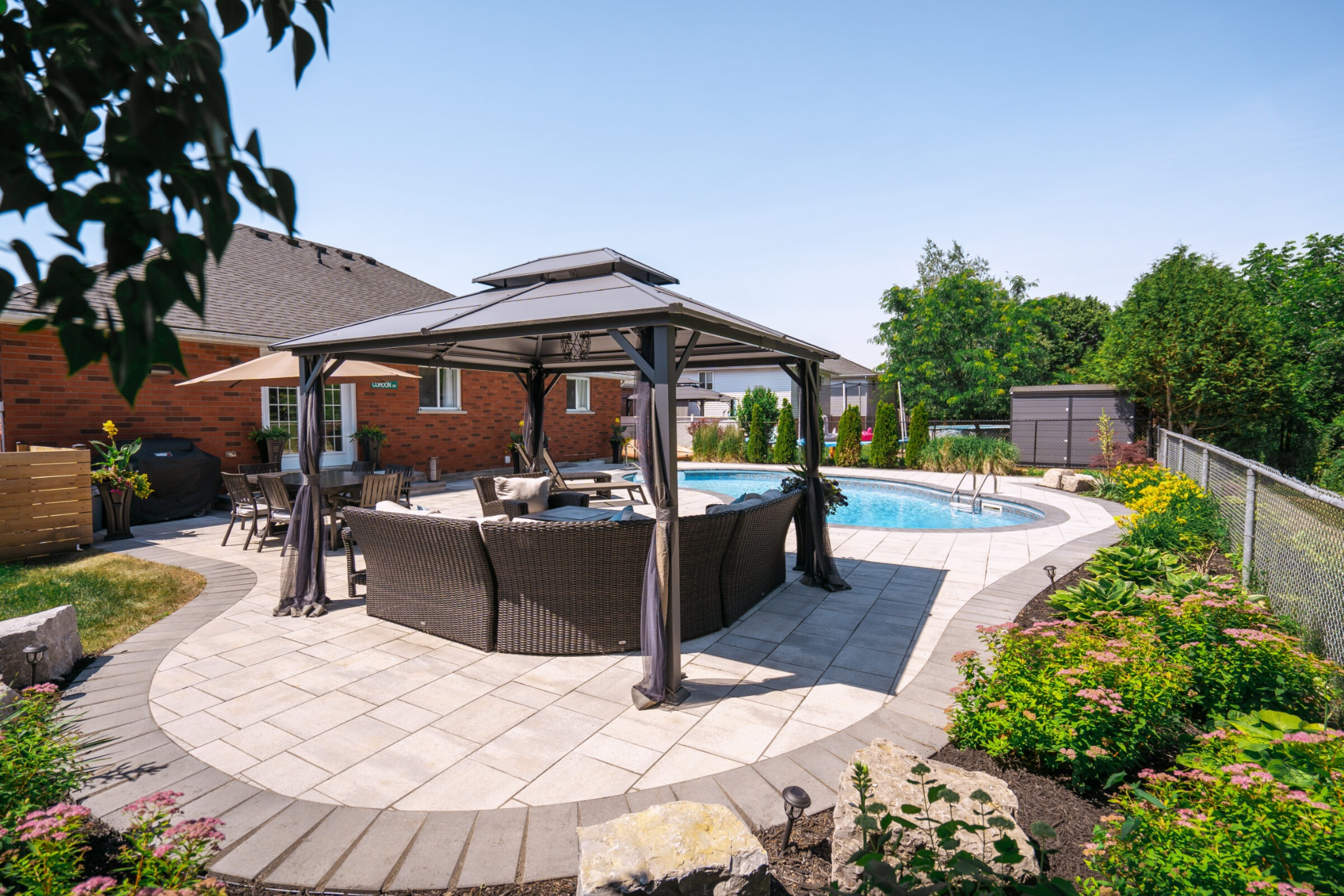 A patio area featuring a dark brown gazebo with a lounge set, situated next to an inground swimming pool. The patio is paved with stone pavers, and there is a brick house in the background.