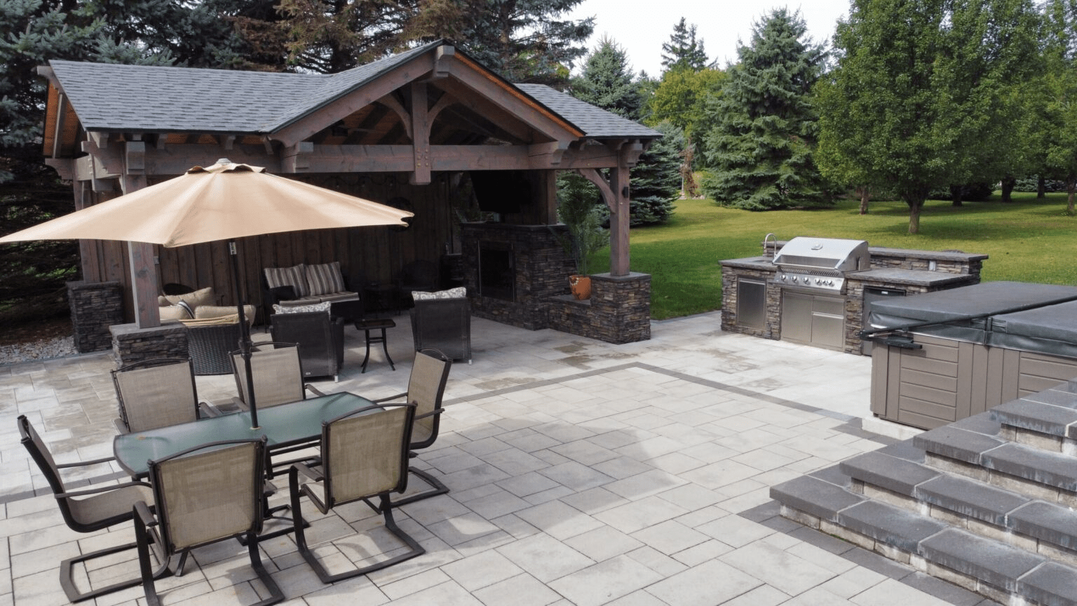 A spacious outdoor living area featuring a wooden pavilion with a stone fireplace, an outdoor kitchen with a stainless steel grill, a dining set with a beige umbrella, and a hot tub on the patio.