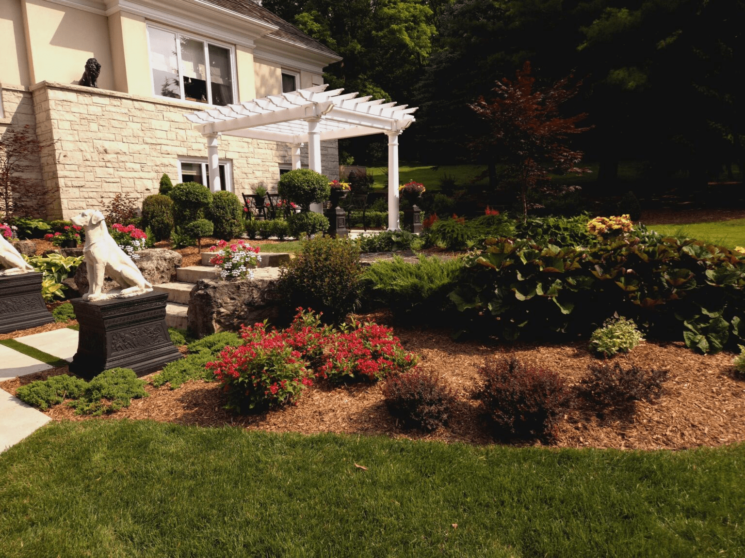 A formal garden scene with a white pergola attached to a stone-walled house. A decorative dog statue sits on a pedestal in the foreground, with manicured shrubs, hostas, and colorful flowering plants in the garden beds.