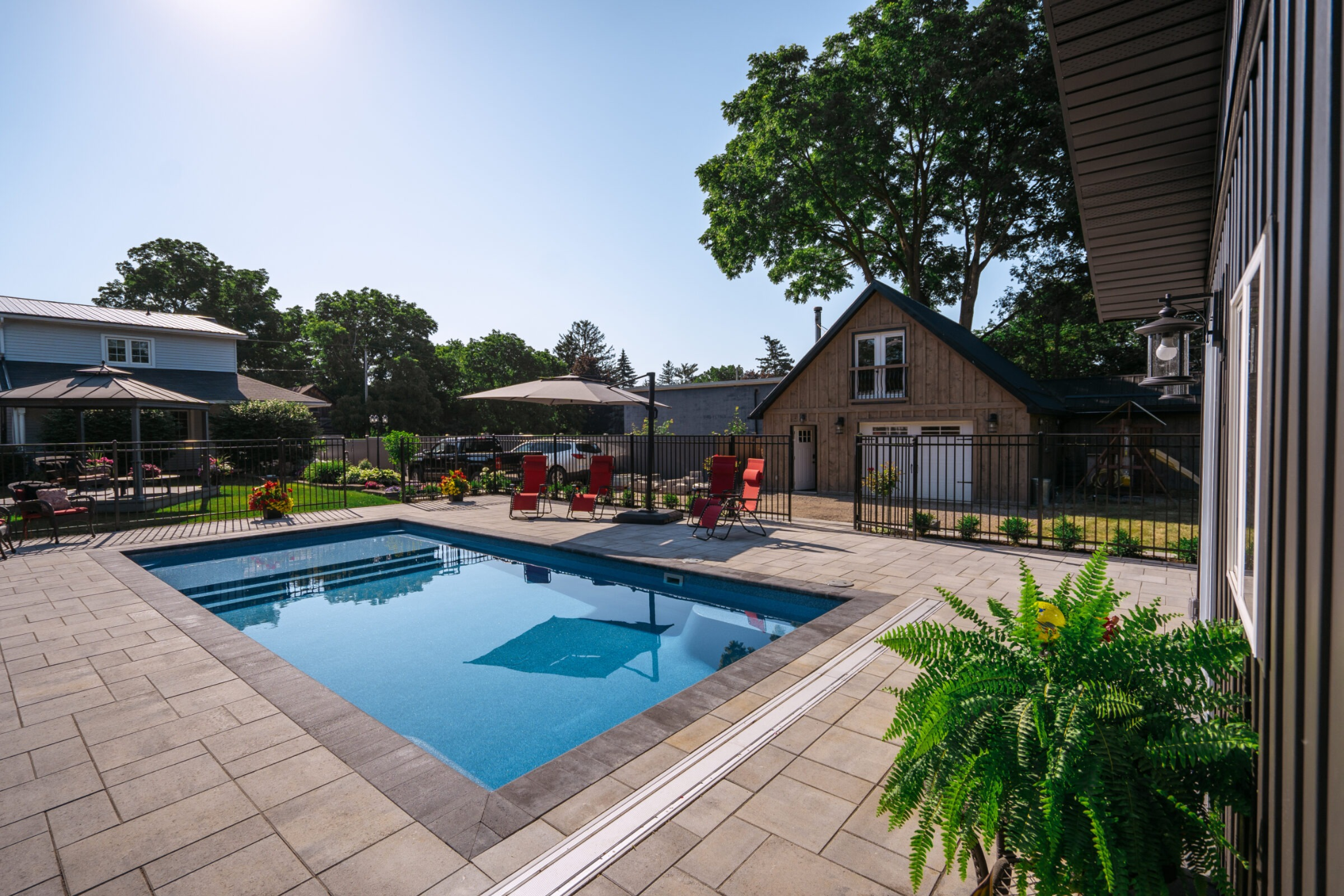 A bright, sunny backyard featuring a rectangular inground pool surrounded by light-colored stone pavers. Red lounge chairs sit poolside under an umbrella, and in the background, there is a rustic-style garage building with a loft and a black metal fence.